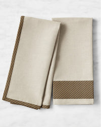 Two beige cloth napkins with black and gold diagonal striped borders, displayed on a white marble surface. One napkin is folded neatly while the other is partially unfolded, showing the border design.