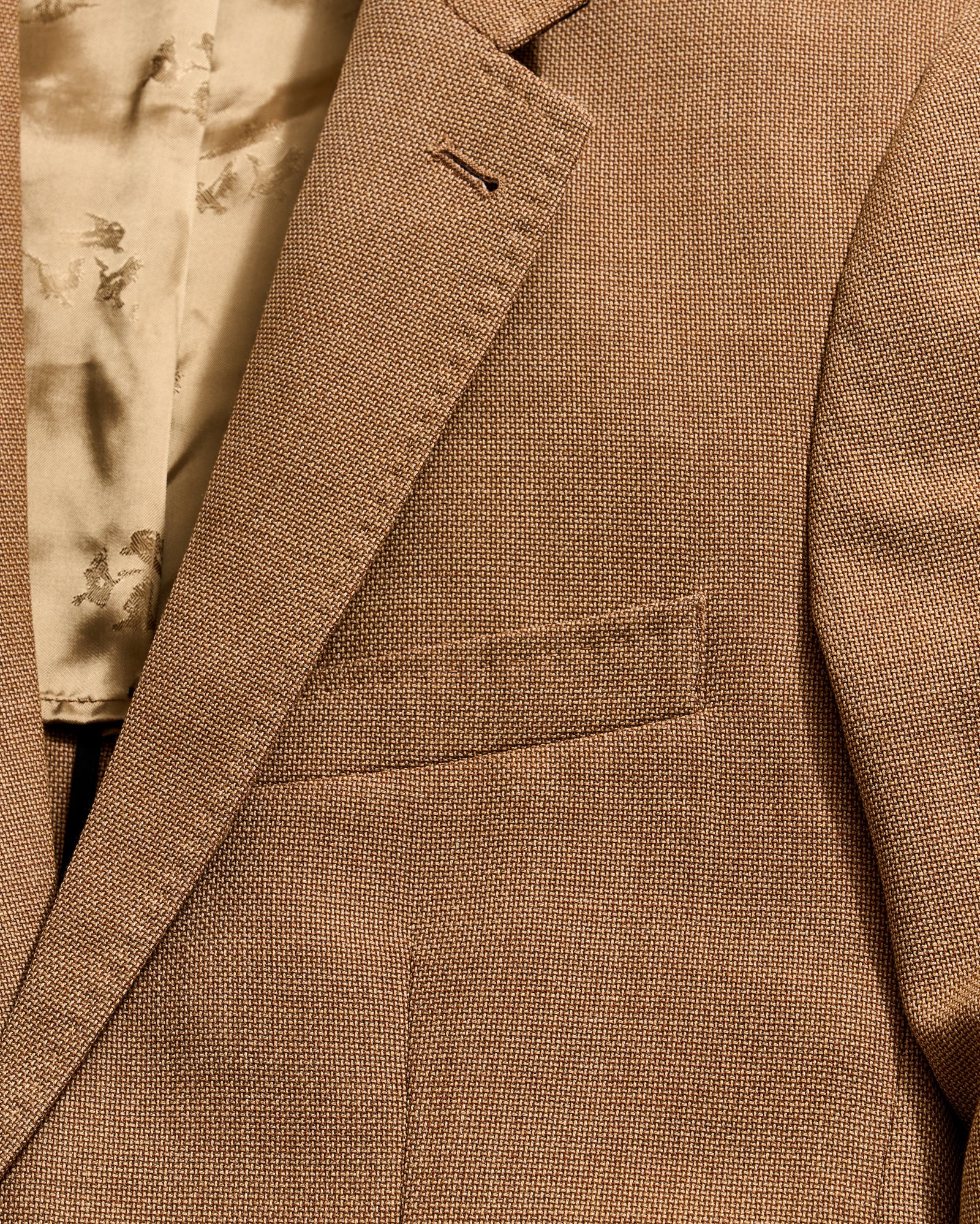 Close-up of a light brown textured suit jacket, featuring a notch lapel, a single buttonhole, and a welt pocket on the chest. The inner lining, visible at the top left, is beige with a subtle pattern.