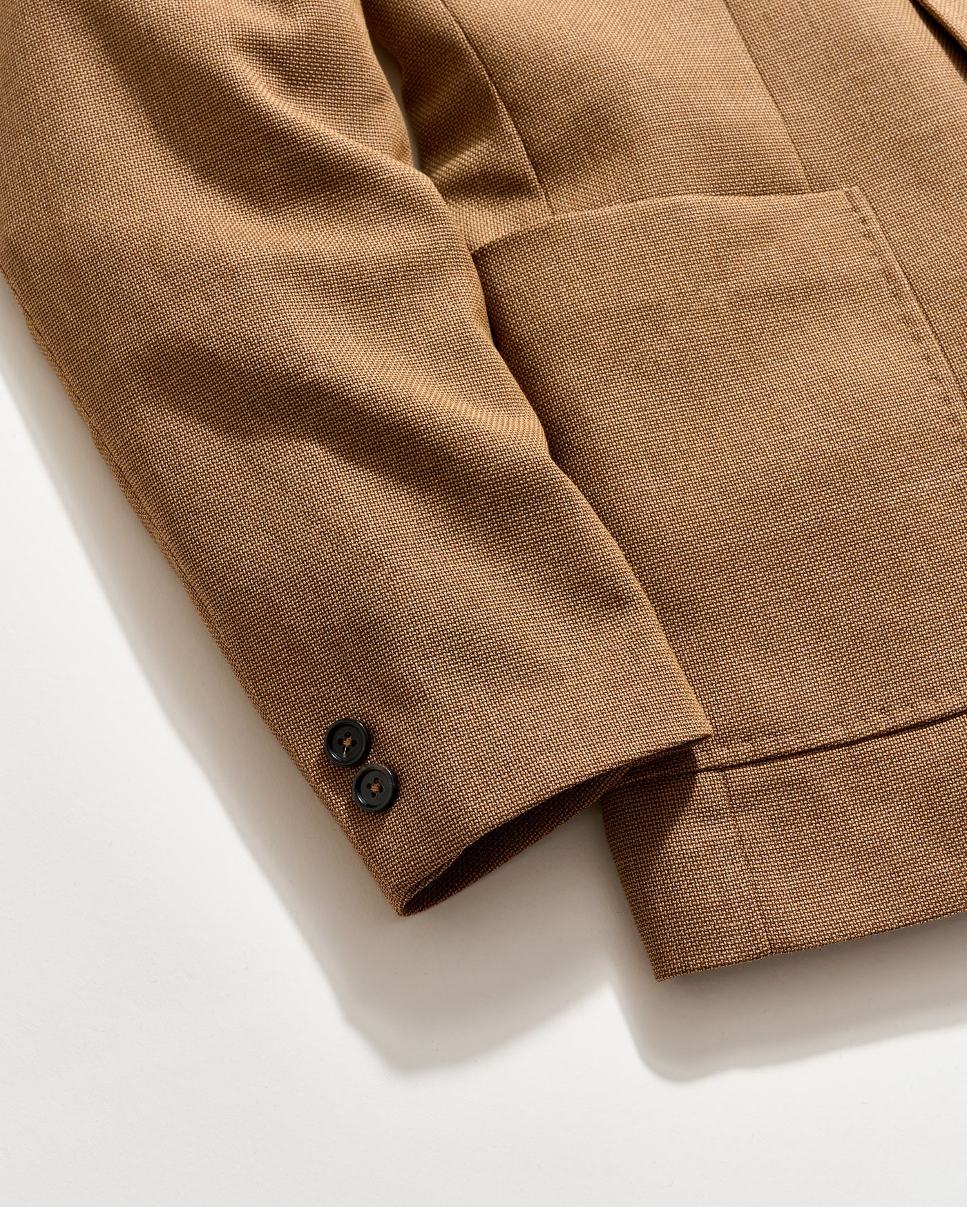 A close-up of a brown textured blazer sleeve with two black buttons on the cuff, shown on a light background. The blazer features a large front pocket.