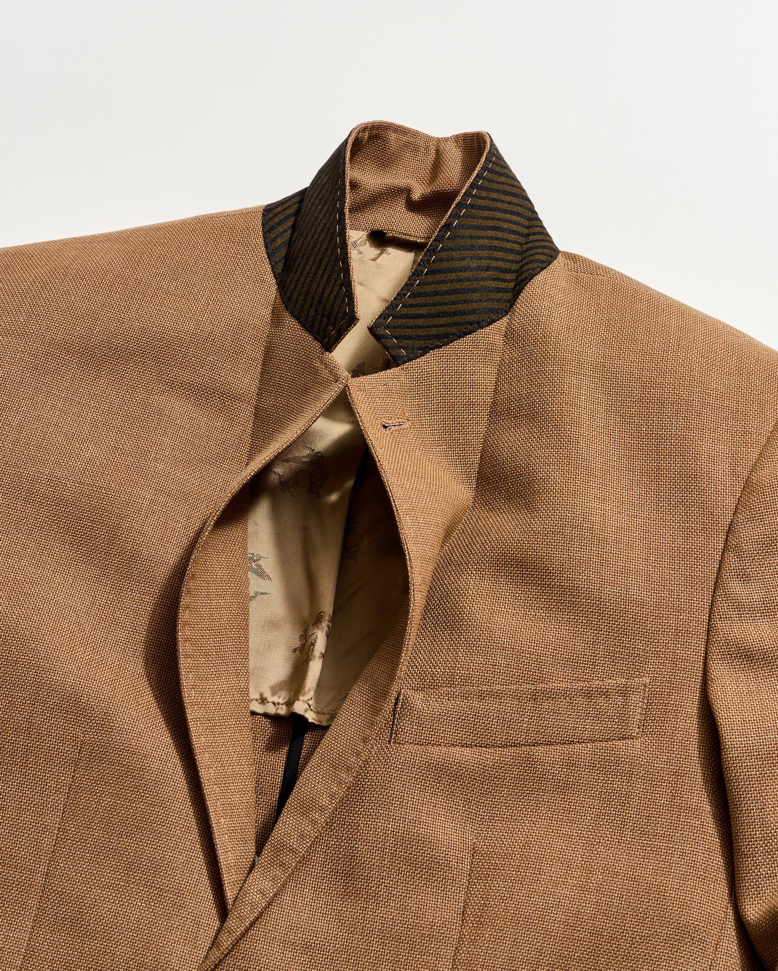 A close-up of a brown suit jacket with a standing collar and a black-striped inner collar. The jacket is partially open, revealing a beige lining with a subtle pattern.