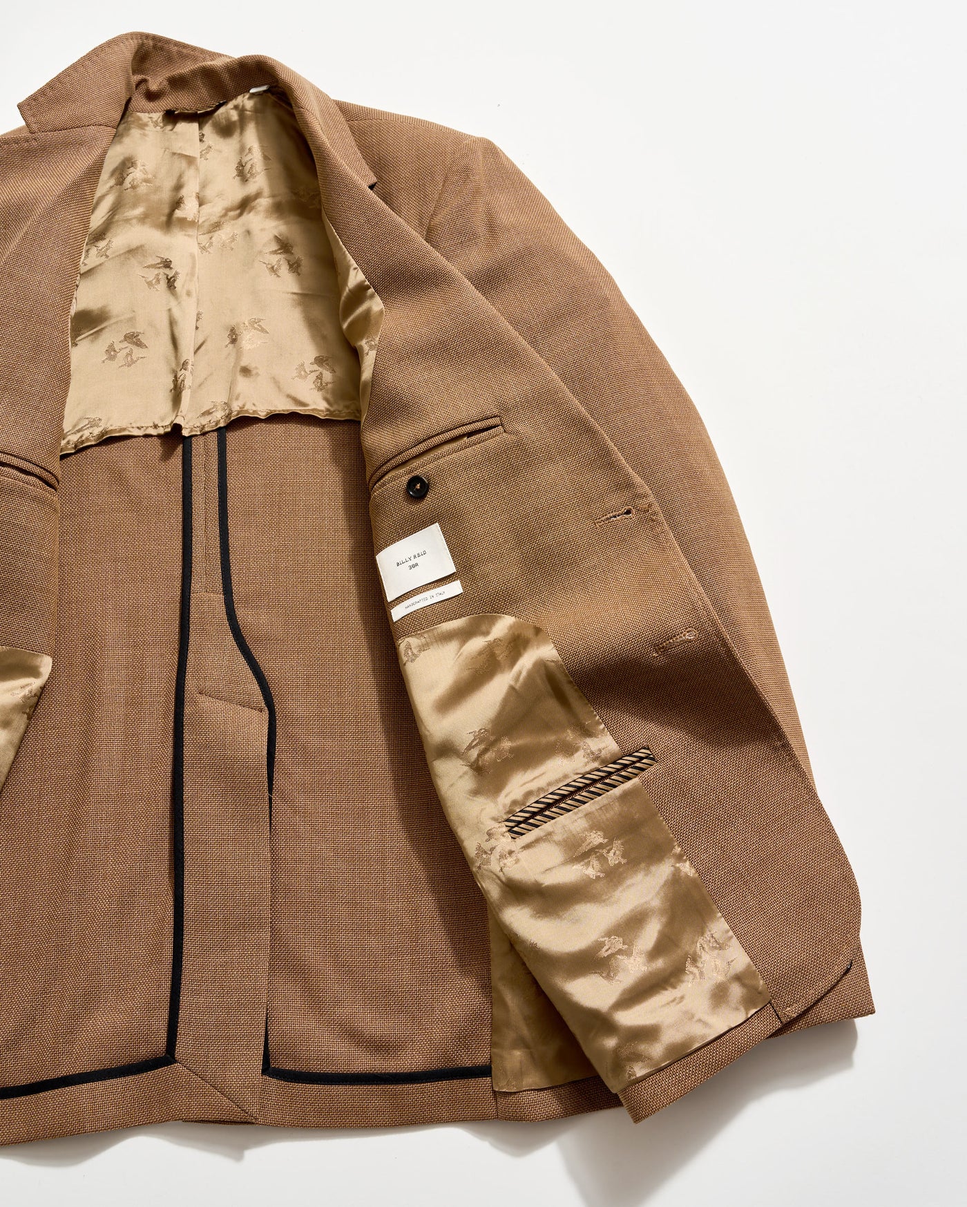 A brown blazer laid open on a white surface, showing its beige, patterned satin lining, multiple interior pockets, a button detail, and a white label sewn inside.