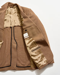 A brown blazer laid open on a white surface, showing its beige, patterned satin lining, multiple interior pockets, a button detail, and a white label sewn inside.
