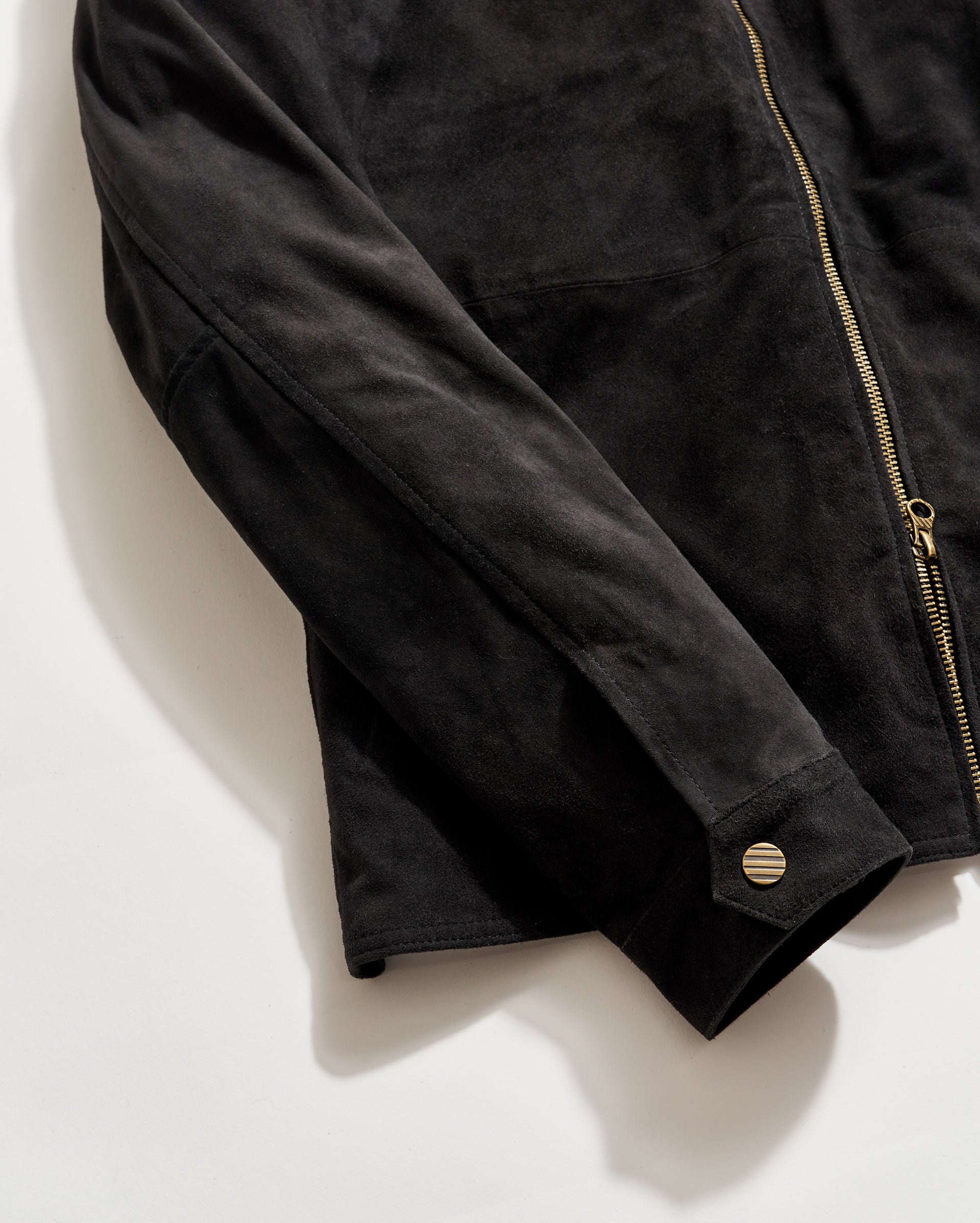 Close-up of a black suede jacket featuring a gold zipper and a snap button on the cuff, displayed against a plain white background.