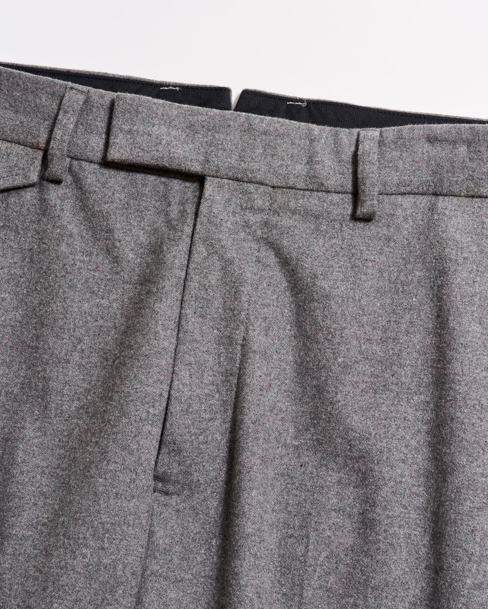 Close-up of the waistband and upper section of a pair of grey dress trousers, showing belt loops, pleats, and part of the zipper area against a plain background.