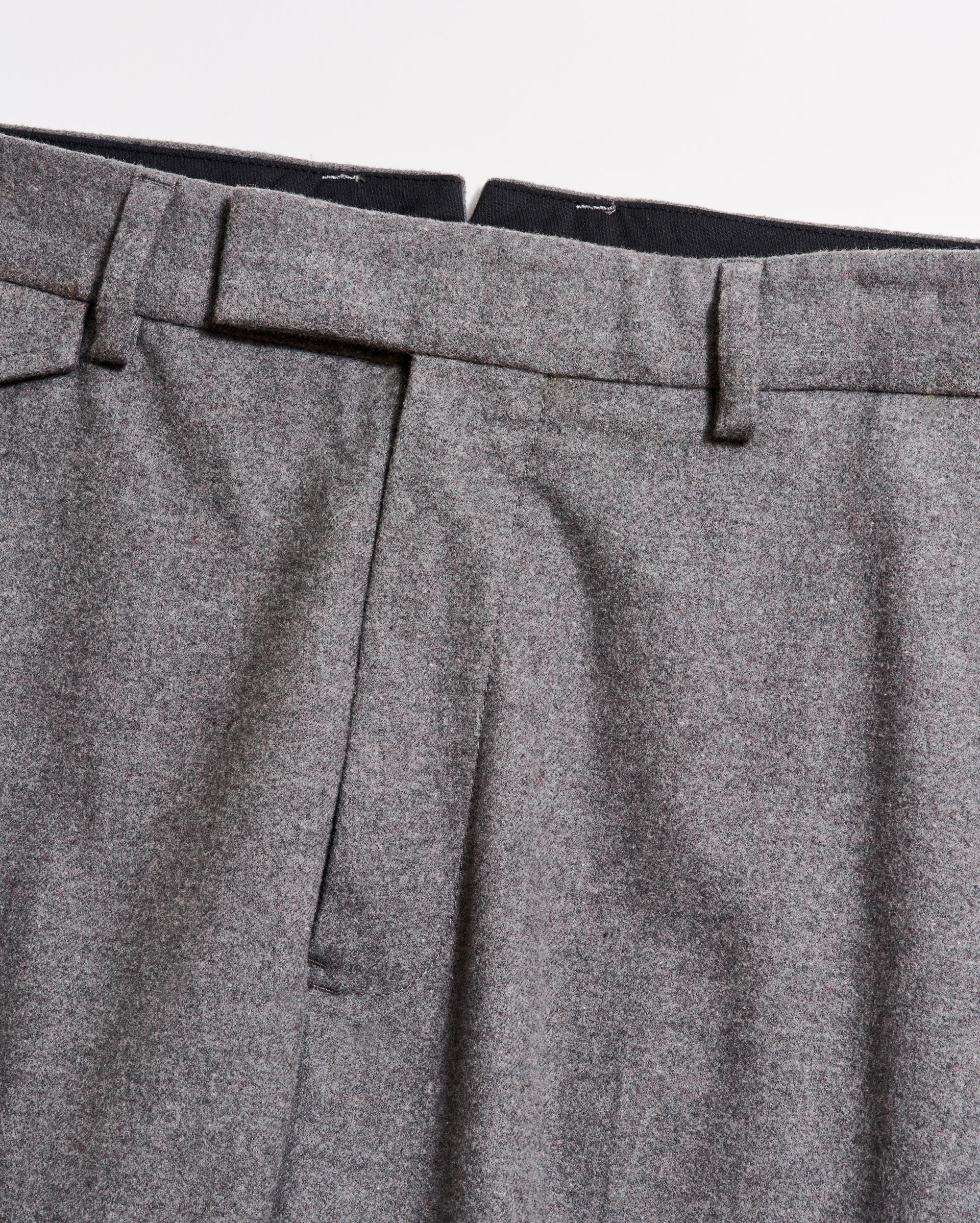 Close-up of the waistband and upper section of a pair of grey dress trousers, showing belt loops, pleats, and part of the zipper area against a plain background.