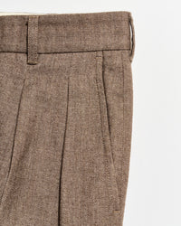 Close-up of brown tweed trousers with pleats, a waistband with belt loops, and a side pocket visible against a white background.