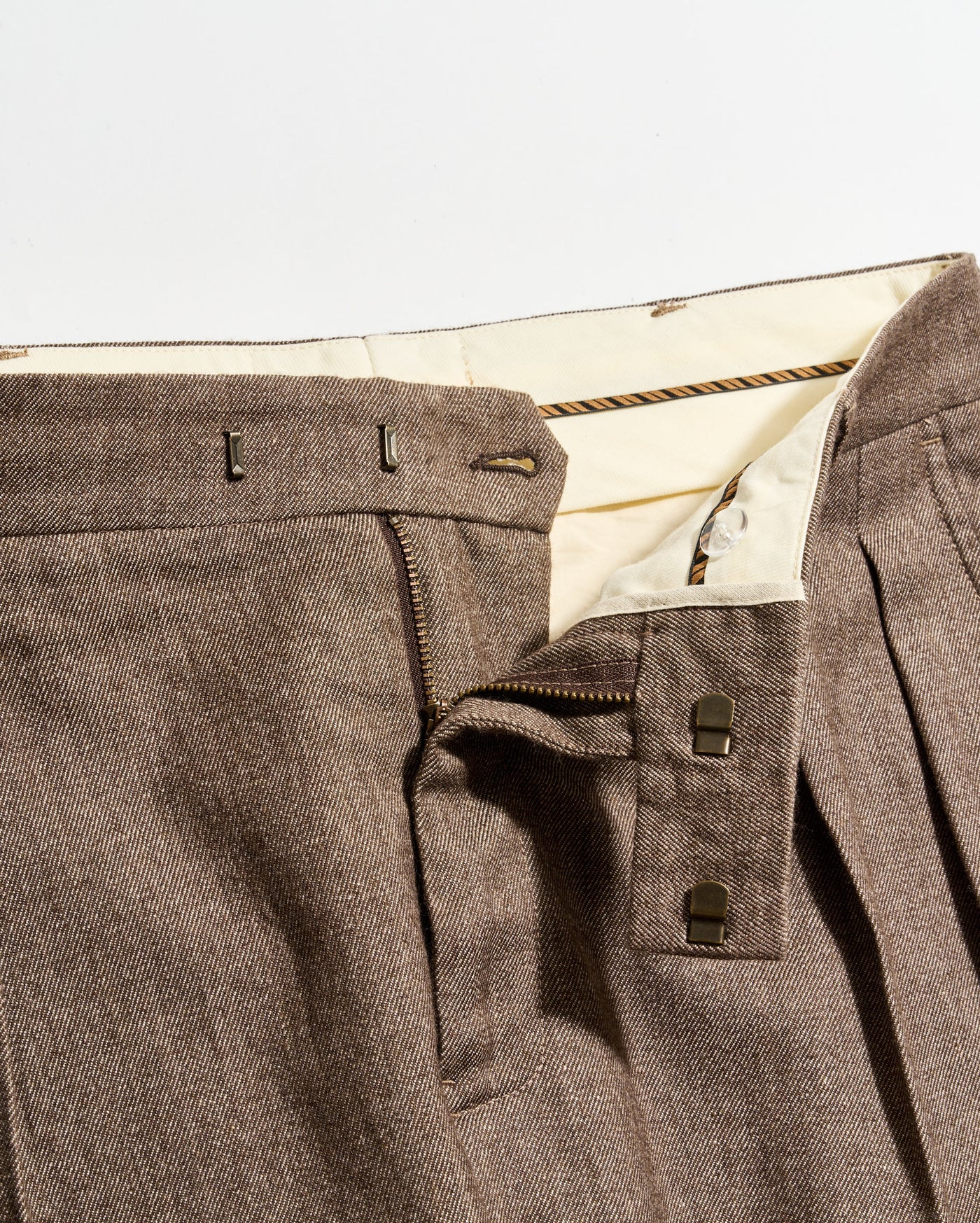 Close-up of brown pleated pants with a partially unzipped fly, showing the inner cream-colored waistband, two metal hook fasteners, and a zipper.