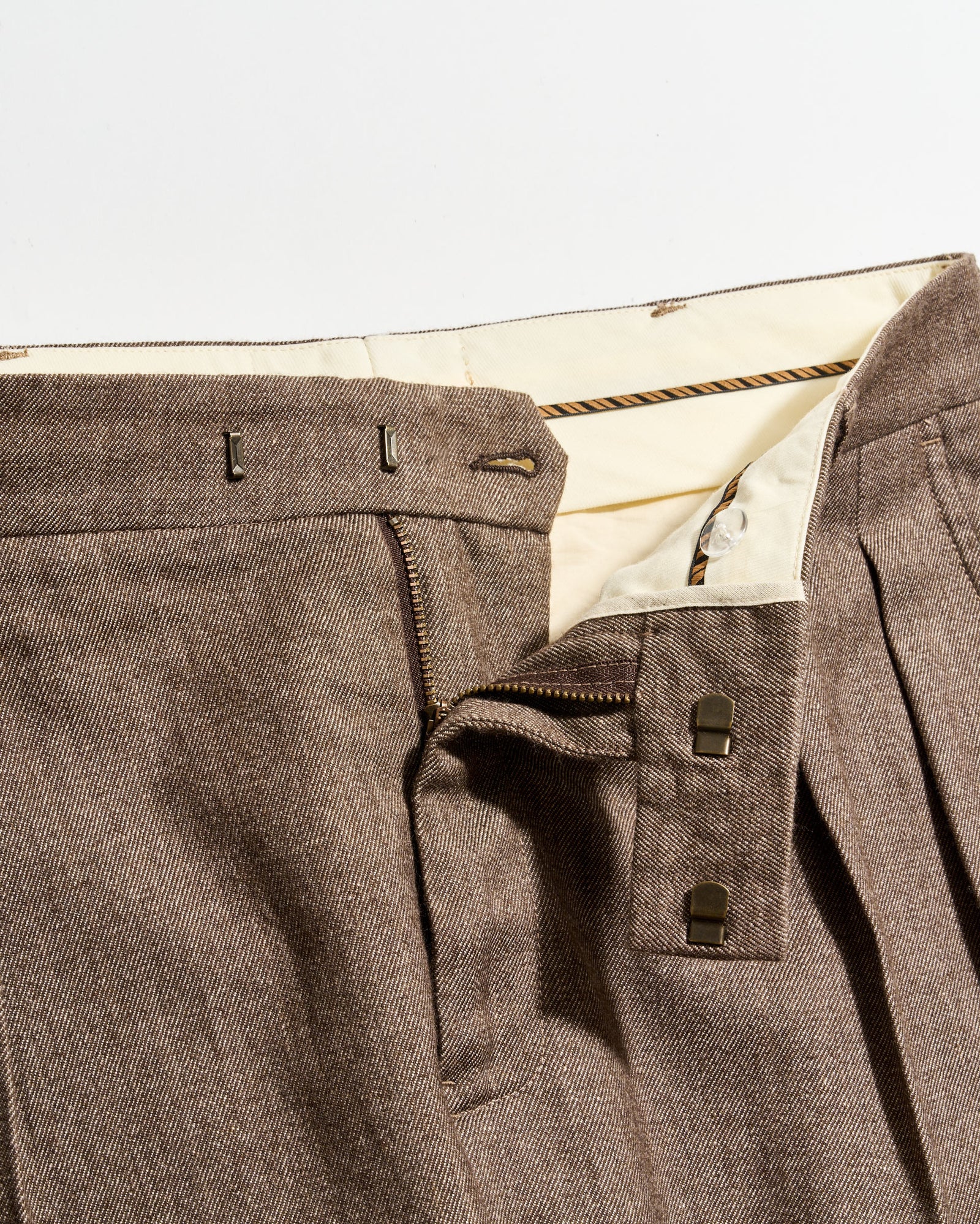 Close-up of brown pleated pants with a partially unzipped fly, showing the inner cream-colored waistband, two metal hook fasteners, and a zipper.