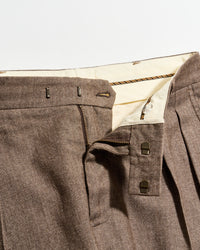 Close-up of brown pleated pants with a partially unzipped fly, showing the inner cream-colored waistband, two metal hook fasteners, and a zipper.