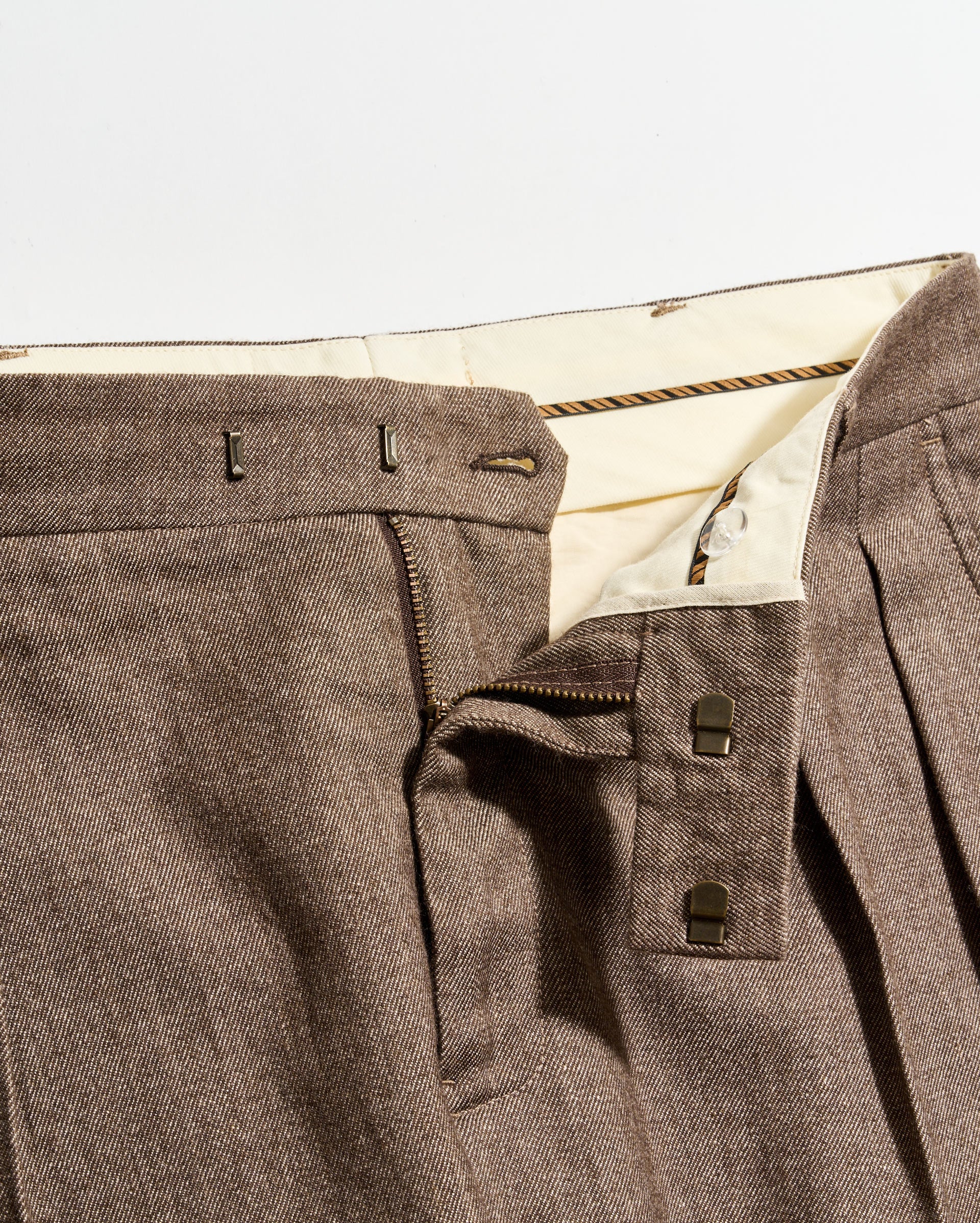 Close-up of brown pleated pants with a partially unzipped fly, showing the inner cream-colored waistband, two metal hook fasteners, and a zipper.