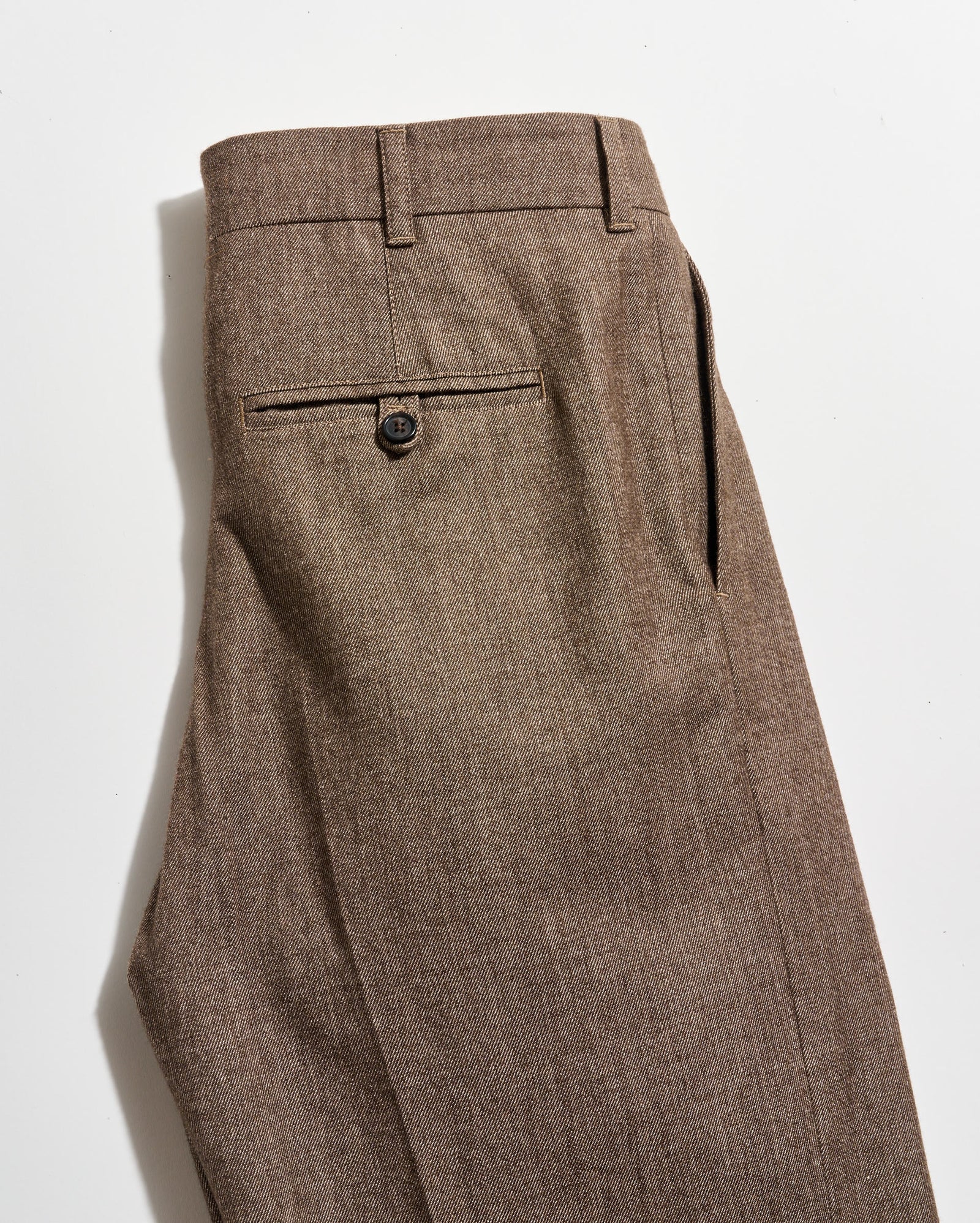 A pair of brown dress pants with a single back pocket featuring a button closure, shown neatly folded on a white background.