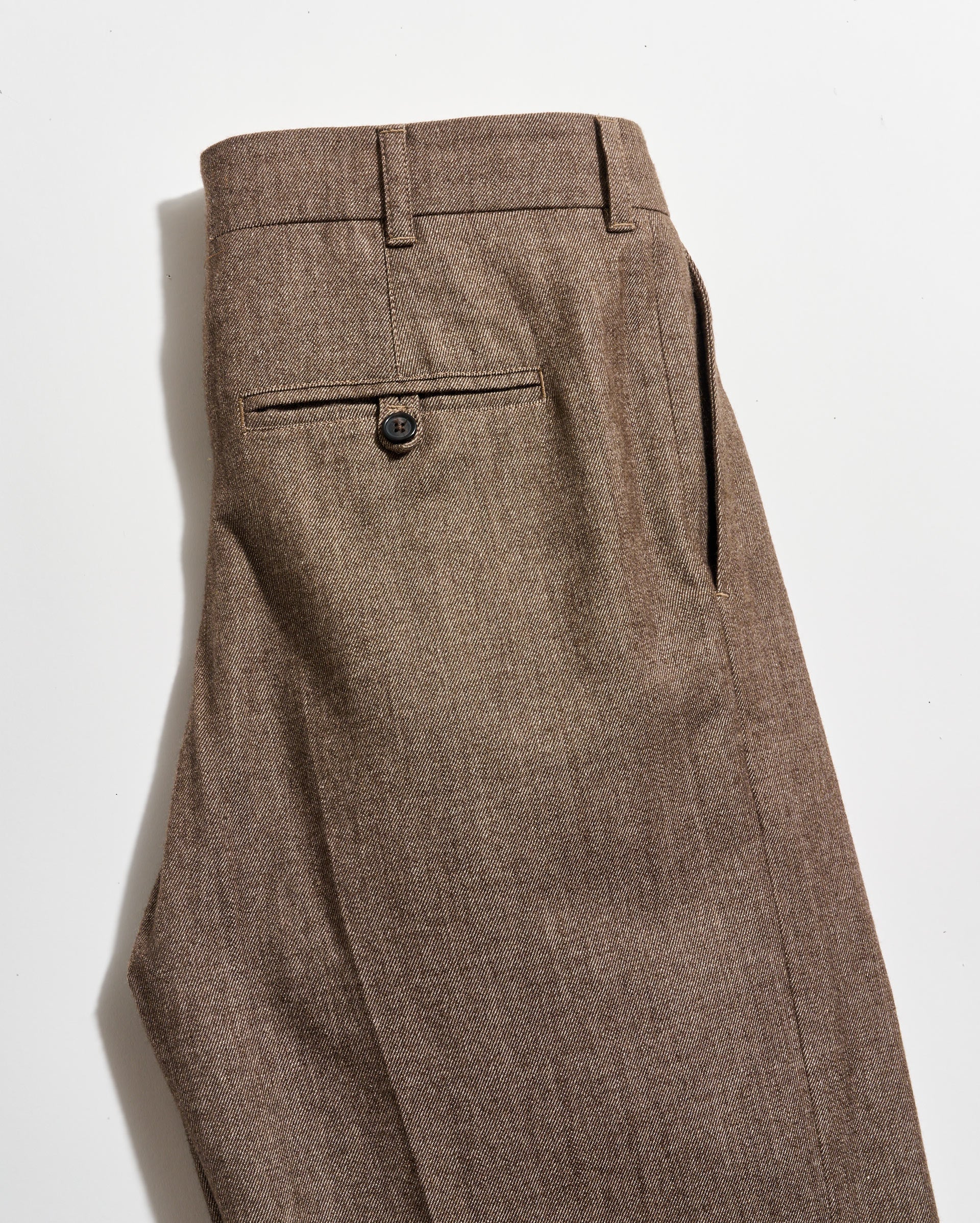 A pair of brown dress pants with a single back pocket featuring a button closure, shown neatly folded on a white background.