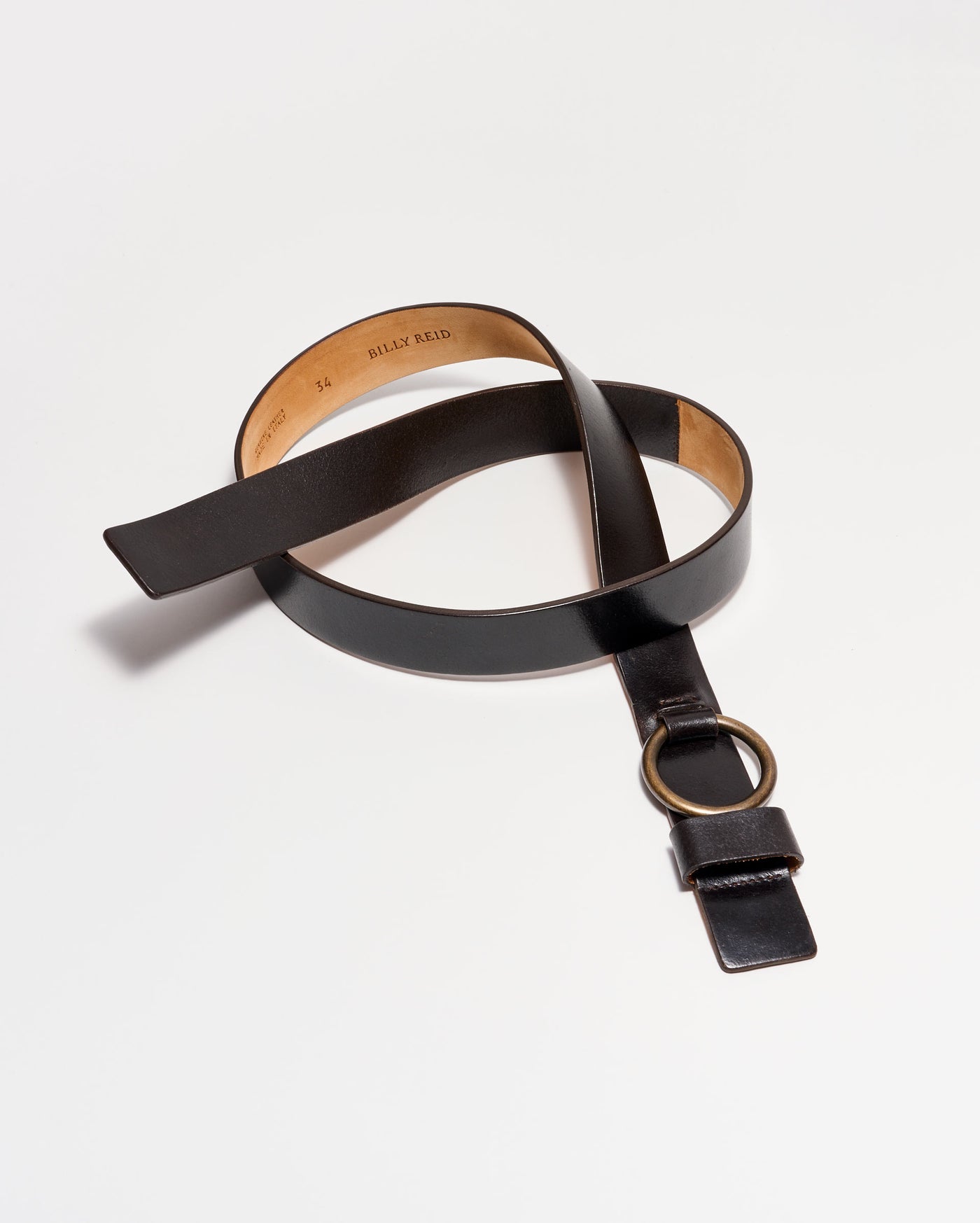 A black leather belt with a brass ring buckle is coiled on a white background. The interior of the belt is a natural tan color and has text stamped on it.