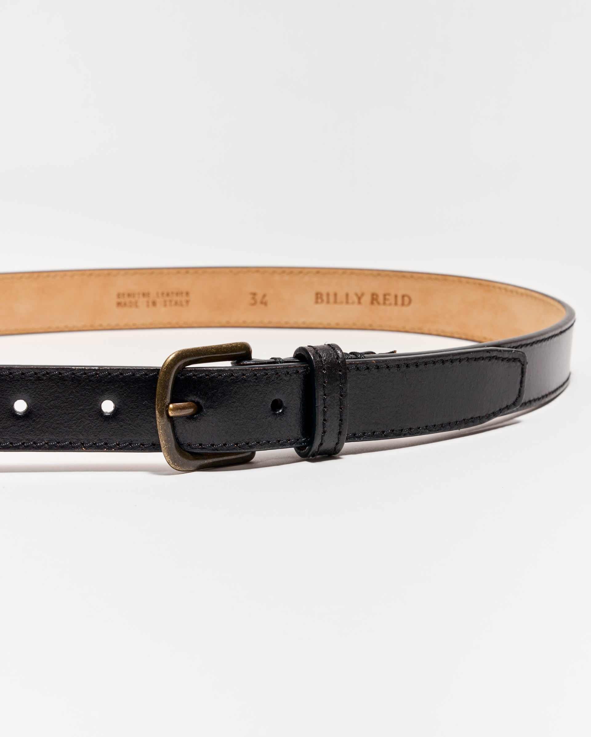 A black leather belt with a brass buckle, shown on a white background. The inside of the belt is tan and stamped with the brand name “Billy Reid,” size 34, and “Genuine Leather Made in Italy.”.