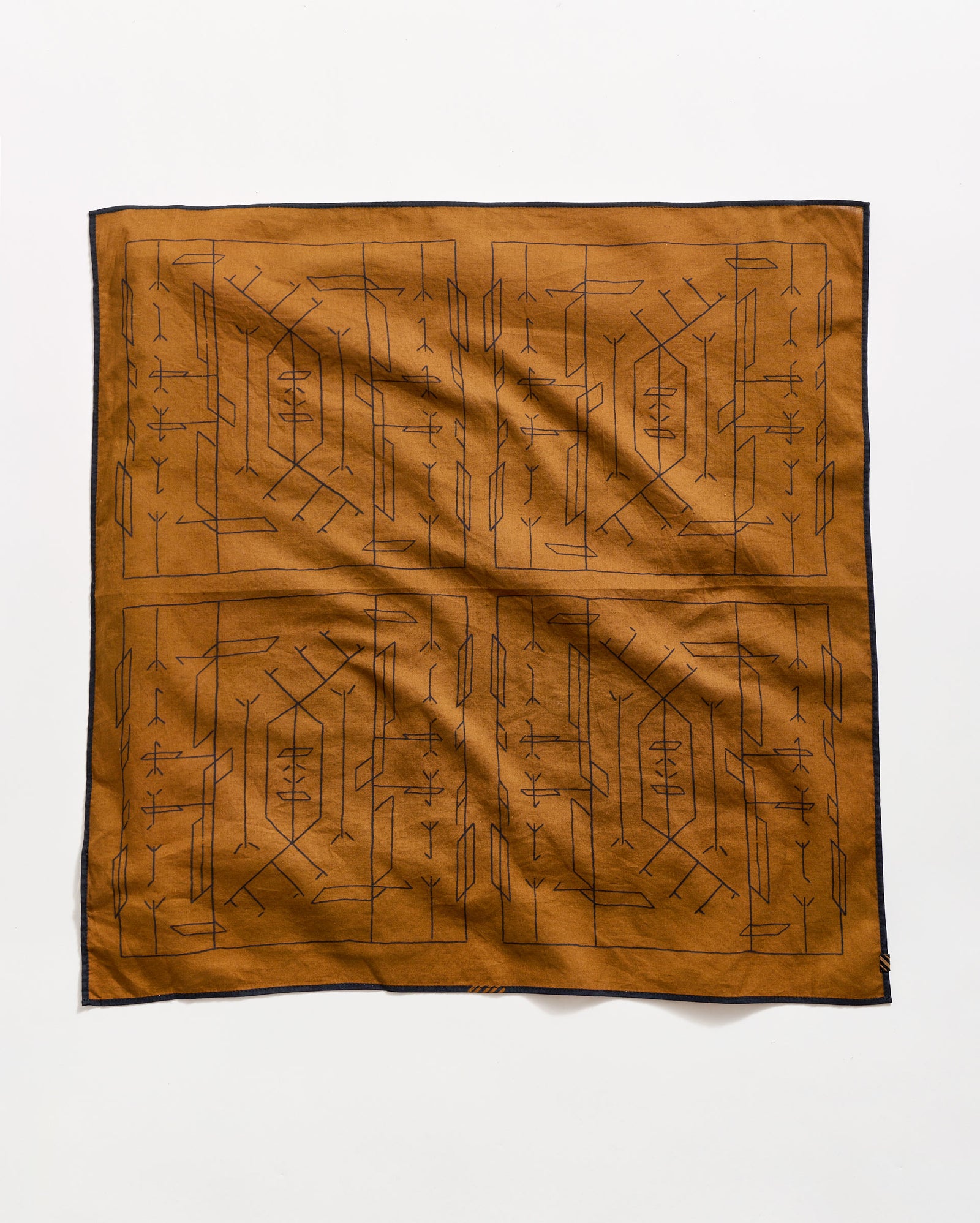 A brown square cloth with a black geometric and linear tribal pattern, bordered by a thin black edge, laid flat on a white background.