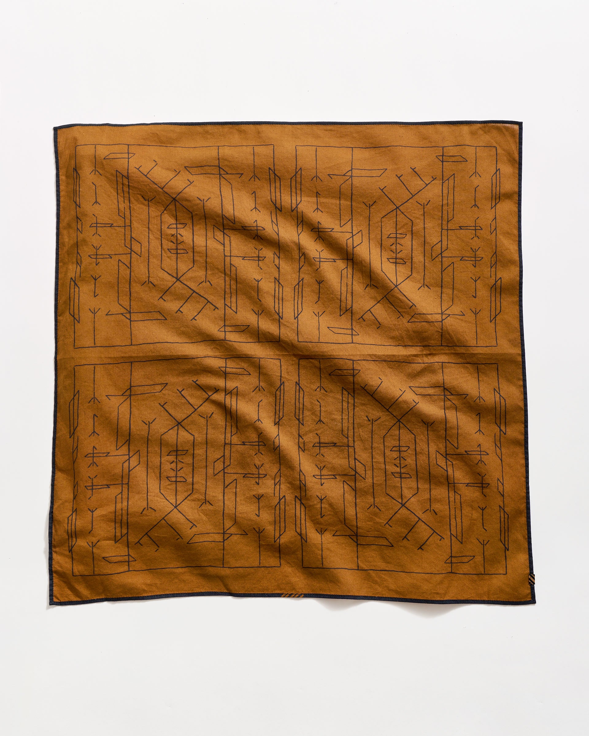 A brown square cloth with a black geometric and linear tribal pattern, bordered by a thin black edge, laid flat on a white background.