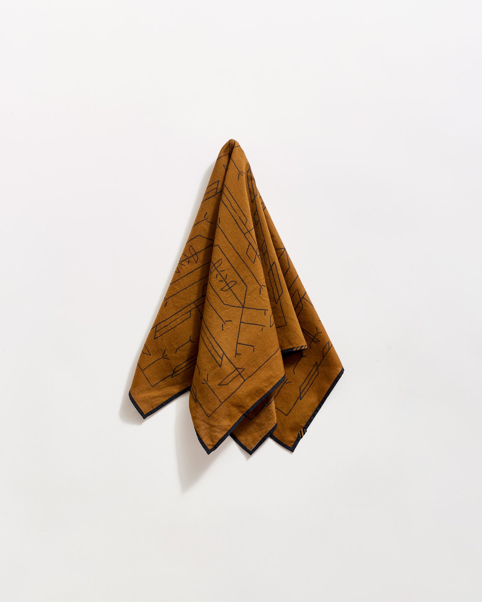 A brown patterned handkerchief or cloth hangs on a white wall, folded at the top and draping downward. The cloth features abstract black line designs and a dark border along the edges.