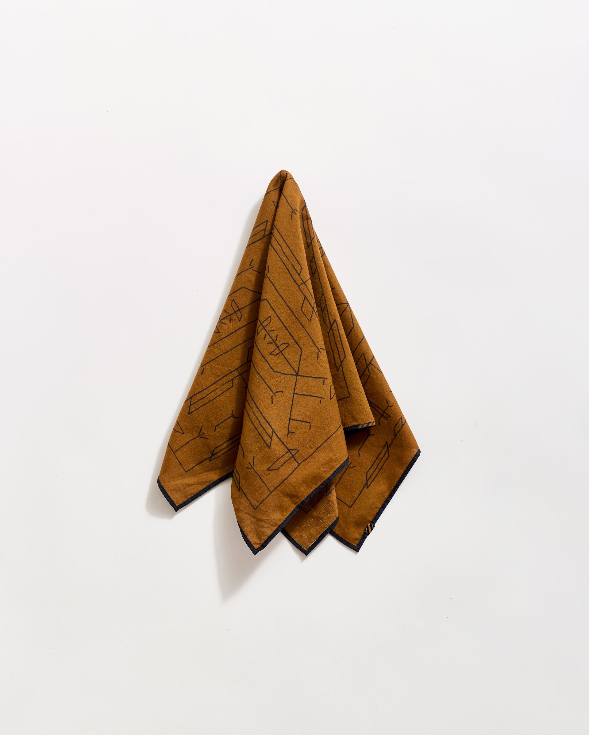 A brown patterned handkerchief or cloth hangs on a white wall, folded at the top and draping downward. The cloth features abstract black line designs and a dark border along the edges.