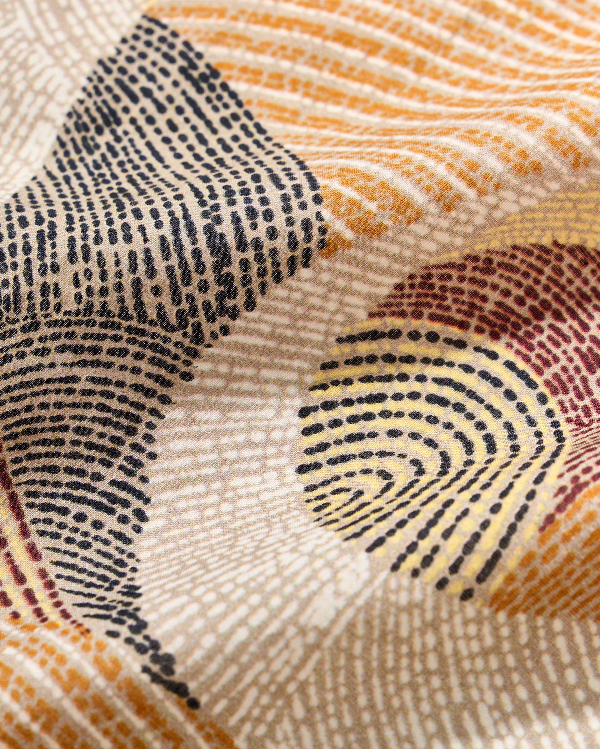 Close-up of fabric with an abstract pattern of concentric dotted lines in black, mustard yellow, beige, and maroon, creating swirling, textured shapes across the cloth surface.