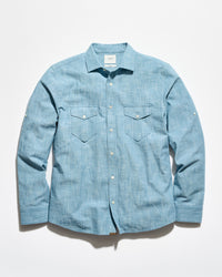A light blue chambray long-sleeve button-up shirt with two chest flap pockets, laid flat against a white background.