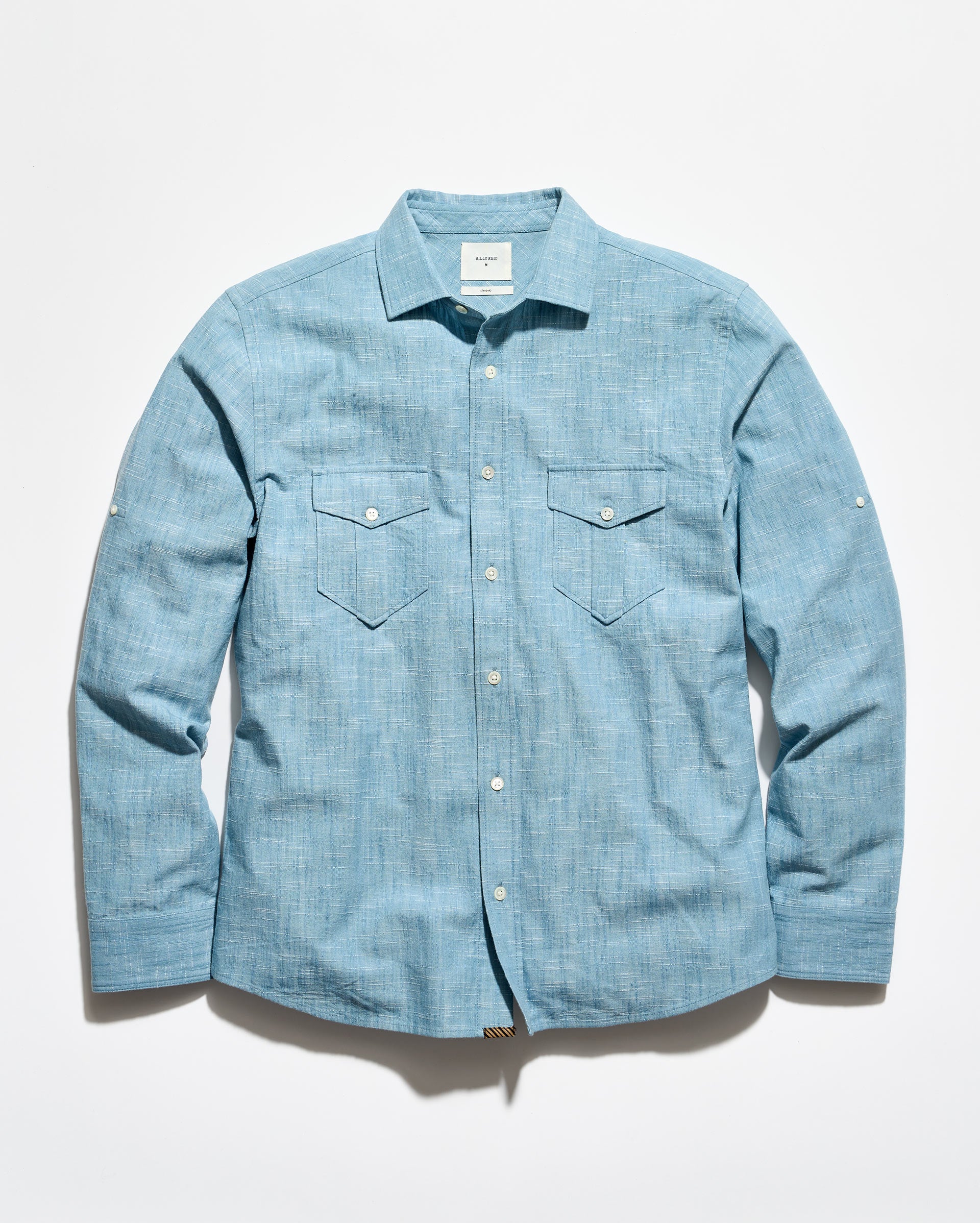 A light blue chambray long-sleeve button-up shirt with two chest flap pockets, laid flat against a white background.