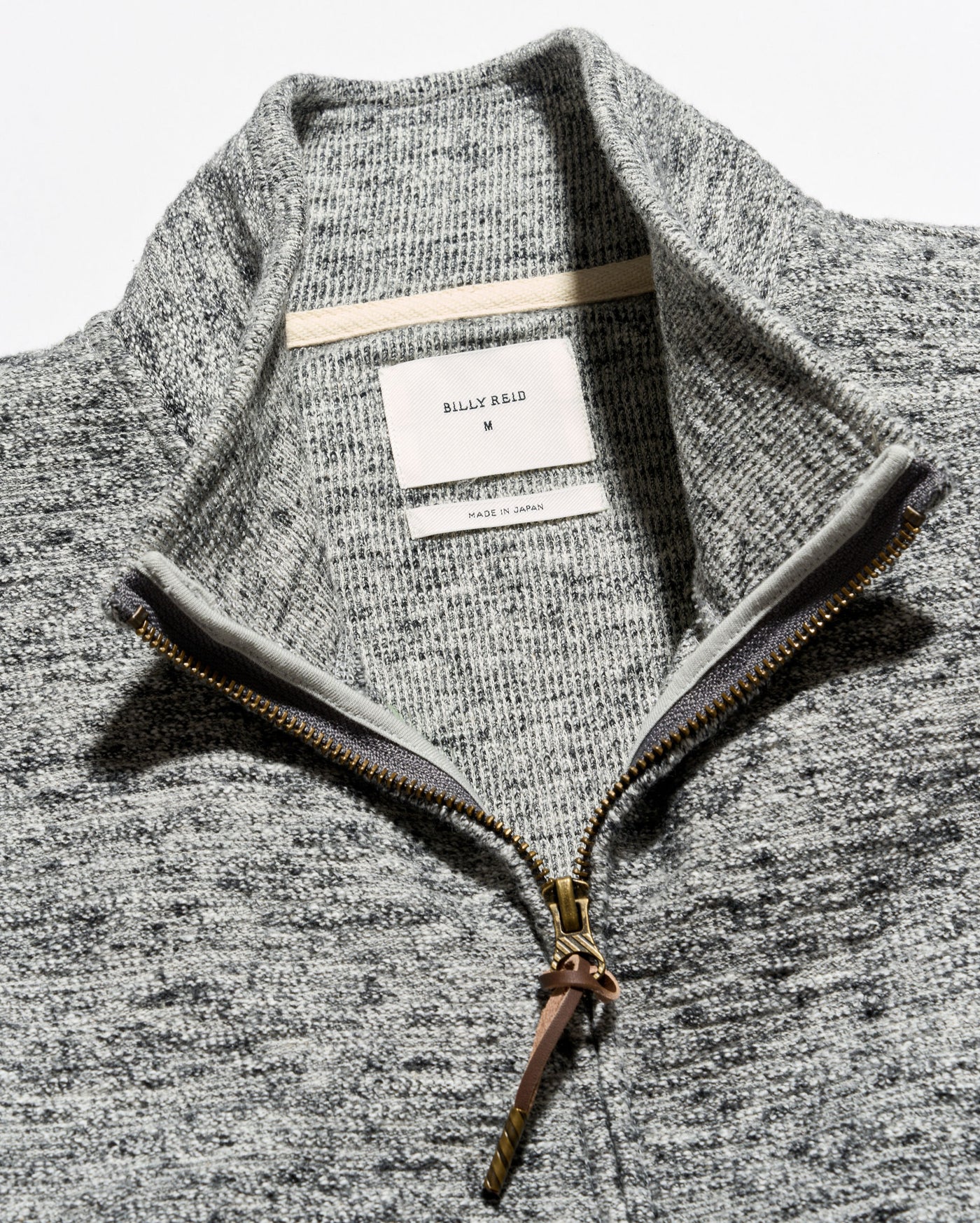 Close-up of a gray heathered knit quarter-zip sweater with a stand collar, showing a white neck label that reads Billie Eilil and a gold-toned zipper.