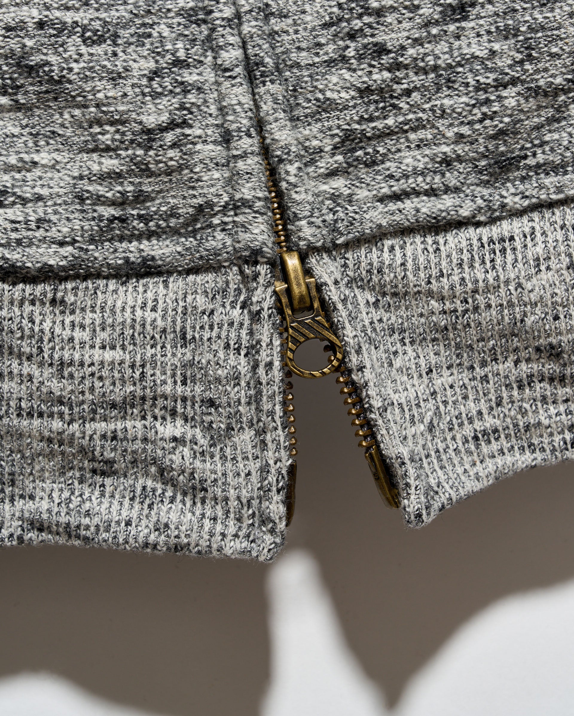 Close-up of a gray and black heathered knit fabric with a gold-tone metal zipper running vertically down the center.