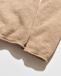 Close-up of a beige knit fabric sleeve or garment detail with visible stitching and a ribbed cuff, photographed on a light neutral background.