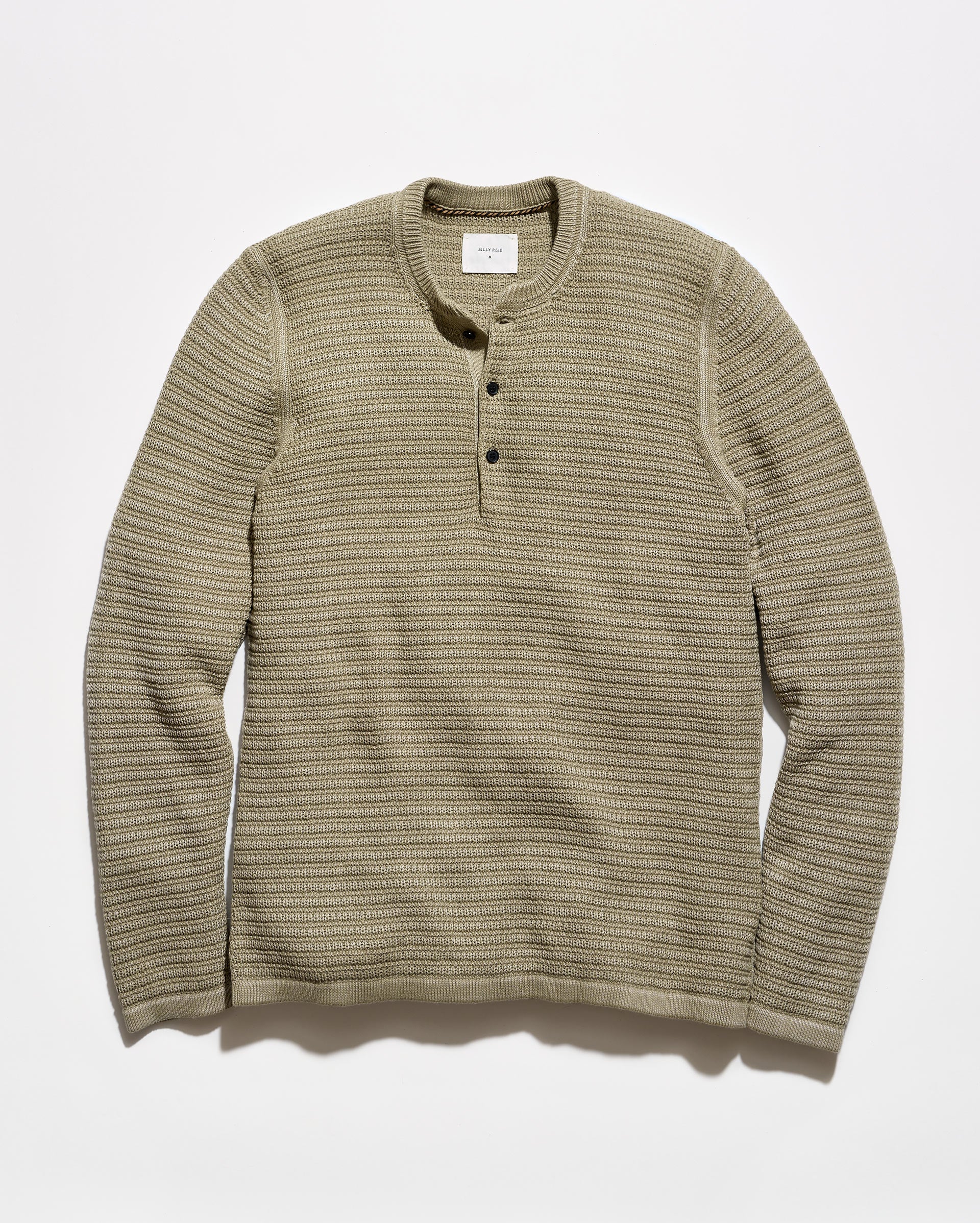 A ribbed knit long-sleeve henley shirt in a muted olive-beige color, featuring a three-button placket and a crew neck, laid flat against a light gray background.