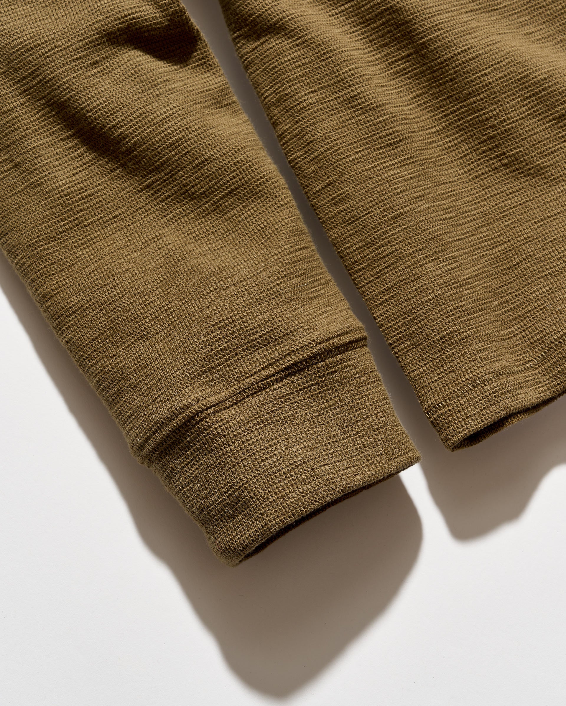 Close-up of a ribbed knit garment sleeve in an olive brown color, laid flat on a white surface with a soft shadow.