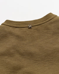 Close-up of a ribbed knit crewneck sweater in olive brown, showing the collar and a small striped detail against a light gray background.