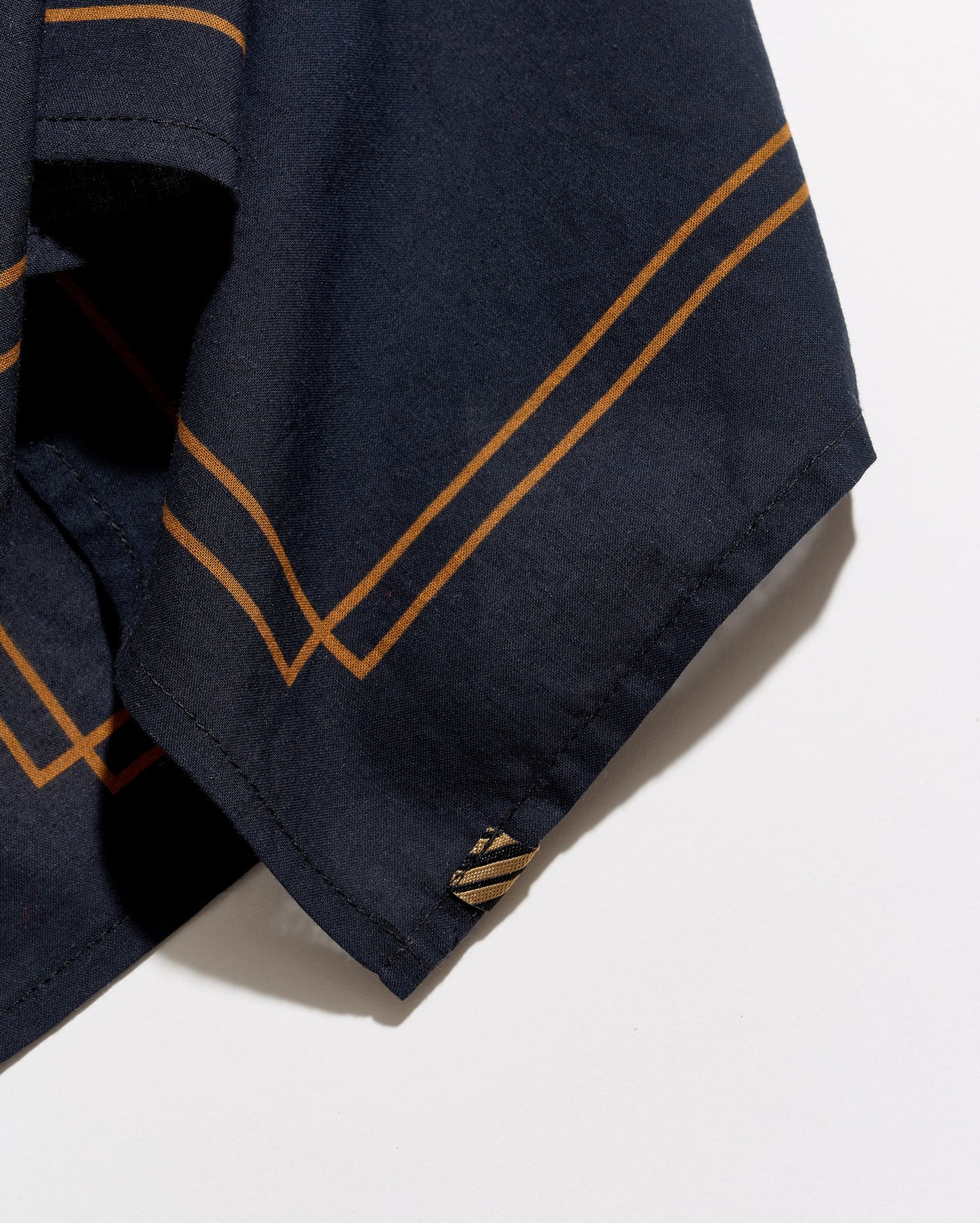 Close-up of a navy blue fabric garment, likely a shirt or jacket sleeve, featuring double diagonal mustard-gold stripes and a small matching embroidered logo near the hem, set against a plain white background.
