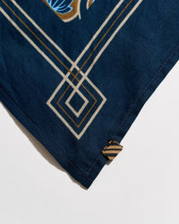 Close-up of a navy blue fabric accessory, likely a scarf or bandana, featuring a geometric diamond pattern in cream and gold, with a small striped fabric tag near the corner.