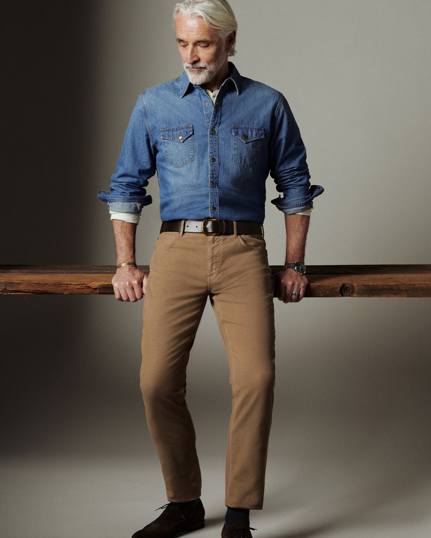 Man wearing a blue denim shirt and beige pants holding a wooden board against a neutral background