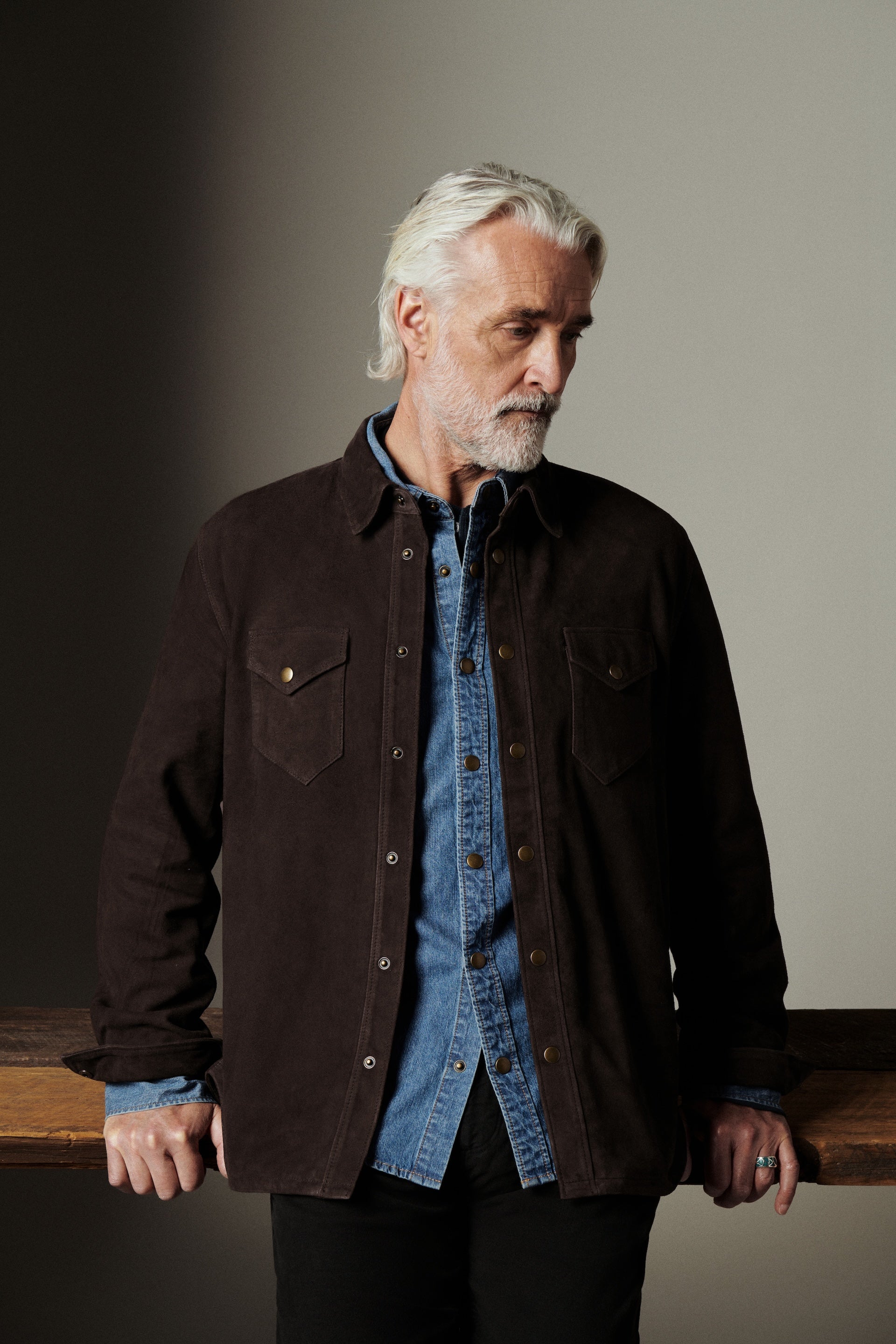 Man wearing a dark jacket over a denim shirt against a neutral background