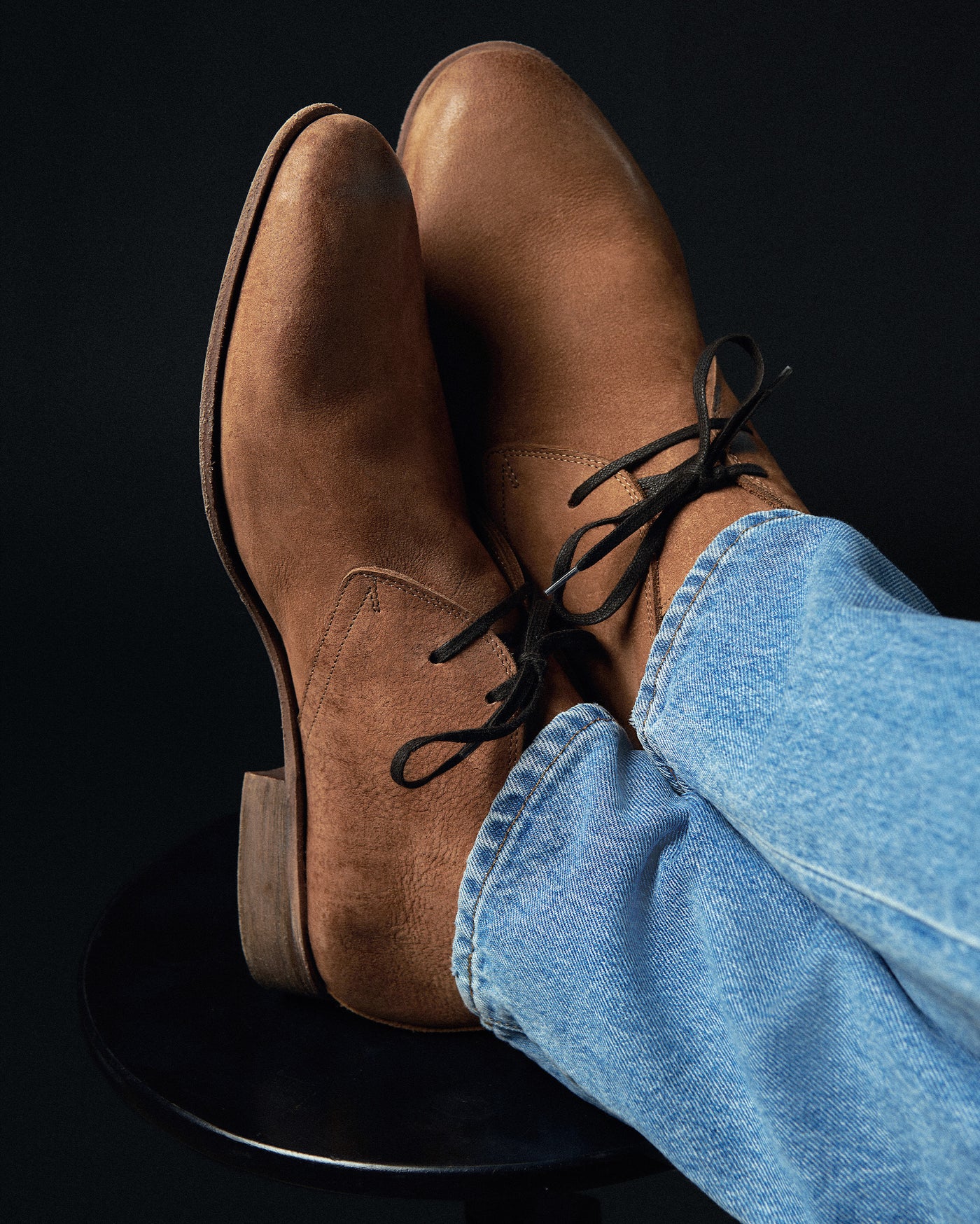Brown leather shoes with black laces worn with blue jeans on a black background