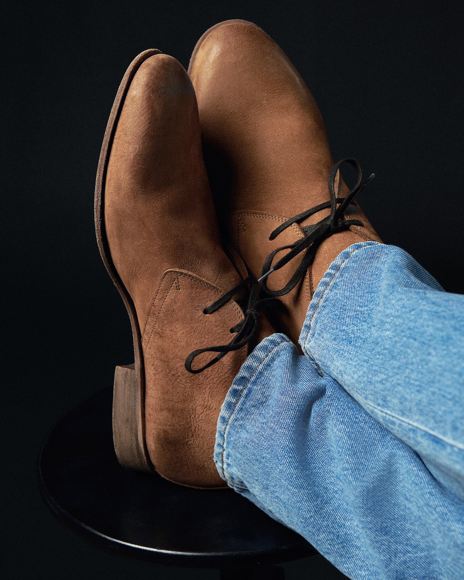 Brown leather shoes with black laces worn with blue jeans on a black background