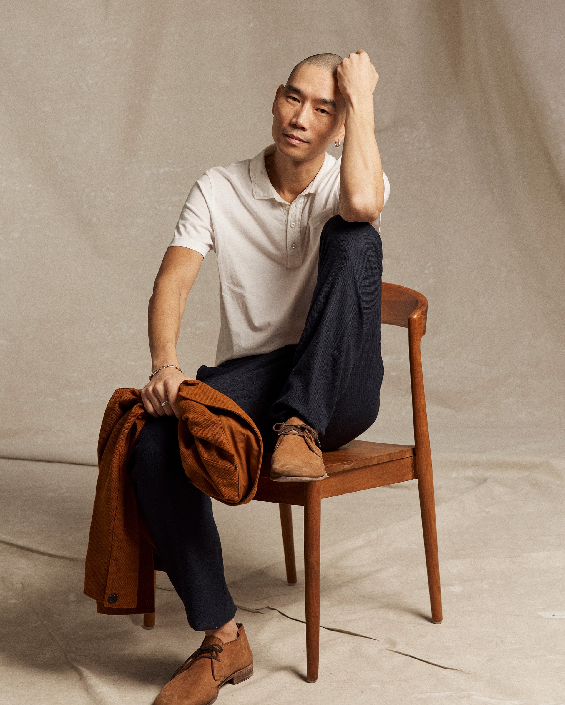 A man with a shaved head sits sideways on a wooden chair, wearing a white polo shirt, dark pants, and brown shoes. He holds a brown jacket in one hand and rests his other arm on his head, against a neutral backdrop.