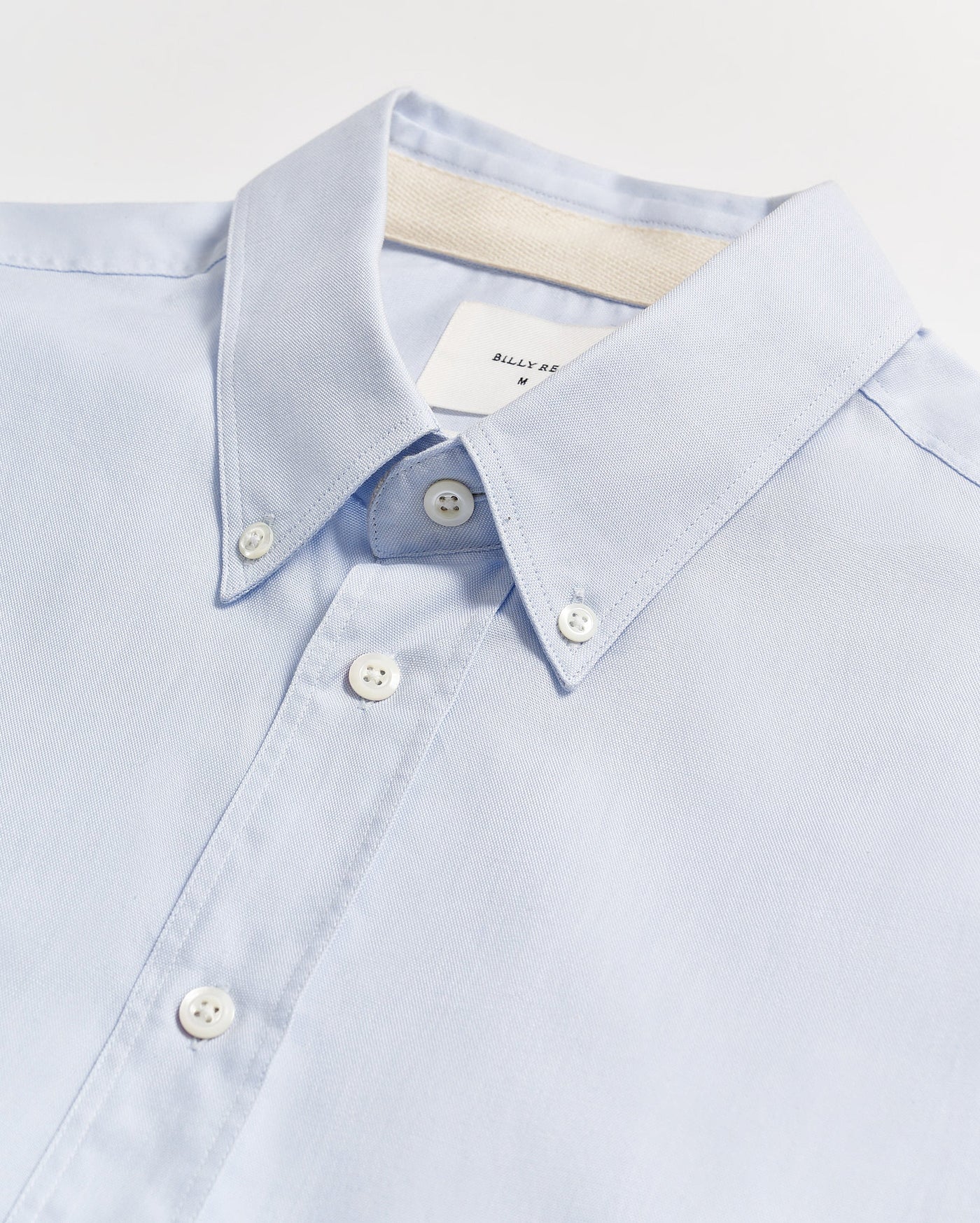 A light blue button-down shirt with white buttons, neatly laid flat on a white surface. The collar is buttoned down, and a white label is partially visible inside the collar.