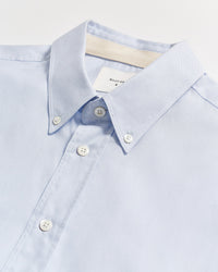 A light blue button-down shirt with white buttons, neatly laid flat on a white surface. The collar is buttoned down, and a white label is partially visible inside the collar.