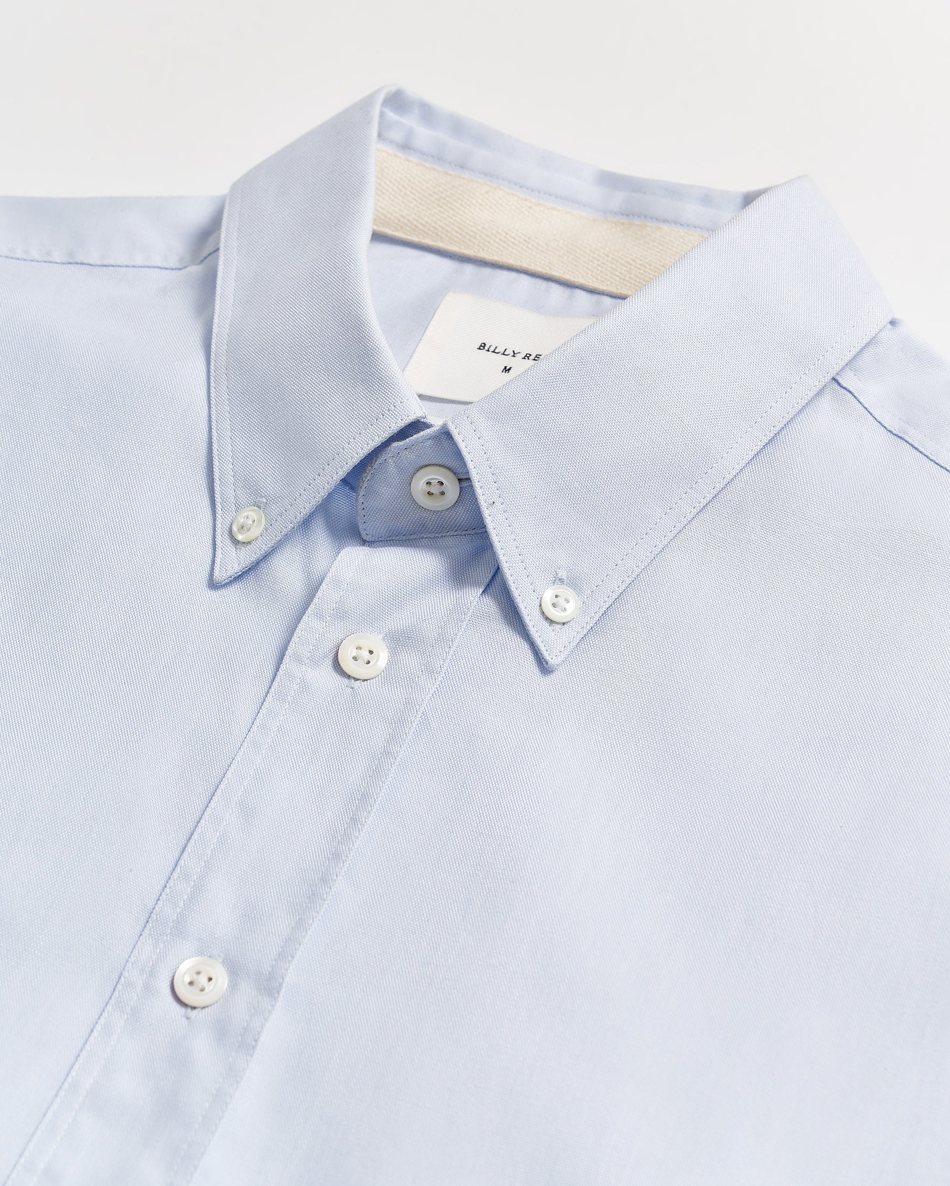 A light blue button-down shirt with white buttons, neatly laid flat on a white surface. The collar is buttoned down, and a white label is partially visible inside the collar.