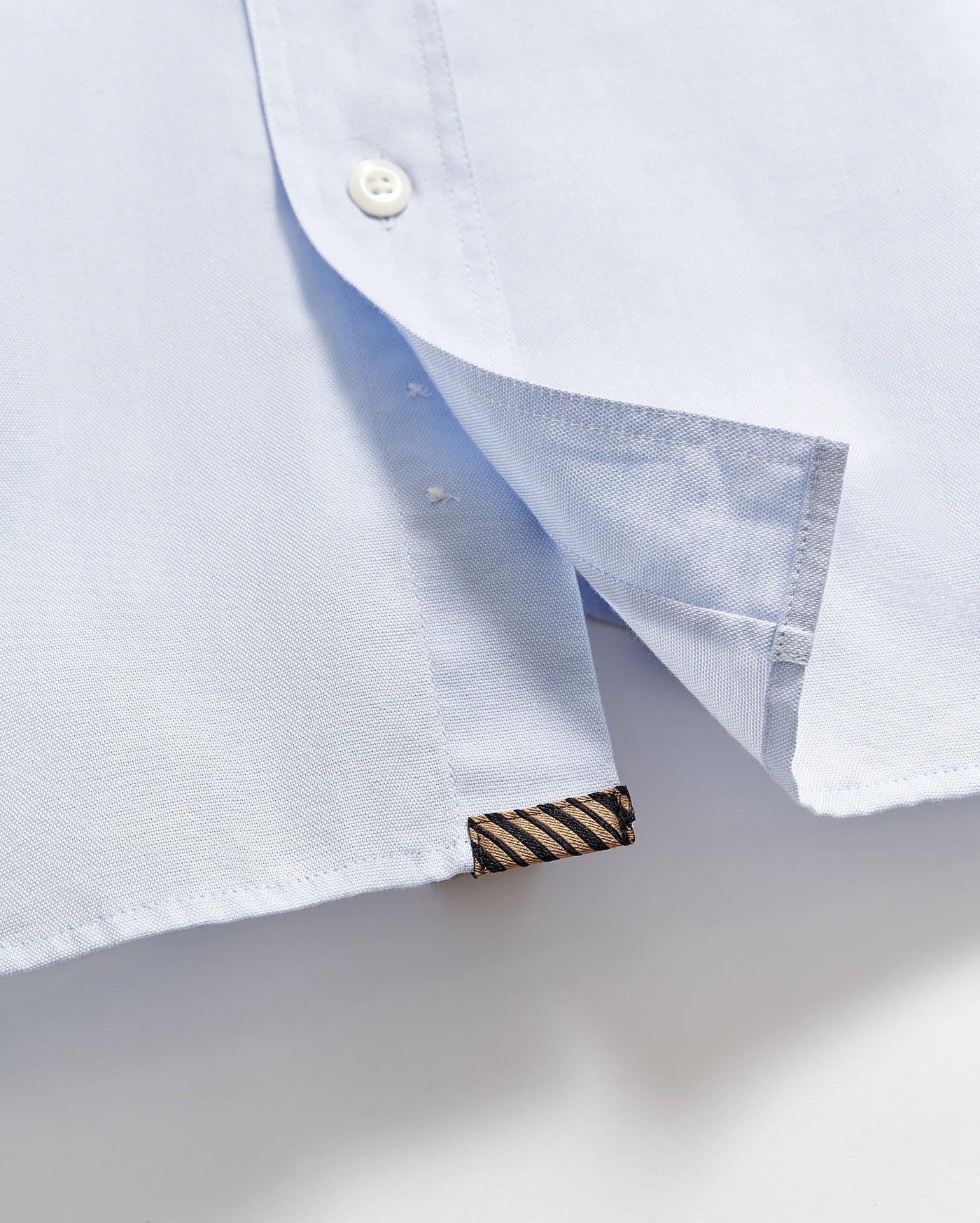 Close-up of the hem of a light blue shirt, showing a white button, a side slit, and a small black and gold striped fabric tag sewn into the bottom edge.