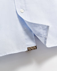 Close-up of the hem of a light blue shirt, showing a white button, a side slit, and a small black and gold striped fabric tag sewn into the bottom edge.