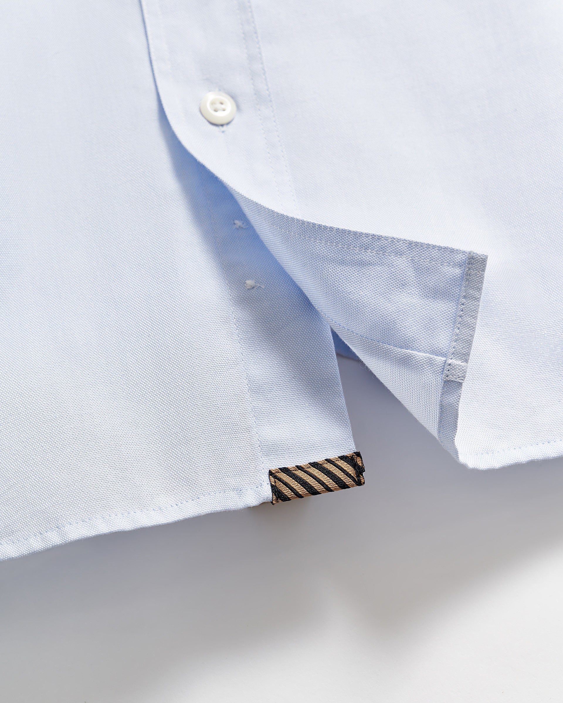 Close-up of the hem of a light blue shirt, showing a white button, a side slit, and a small black and gold striped fabric tag sewn into the bottom edge.