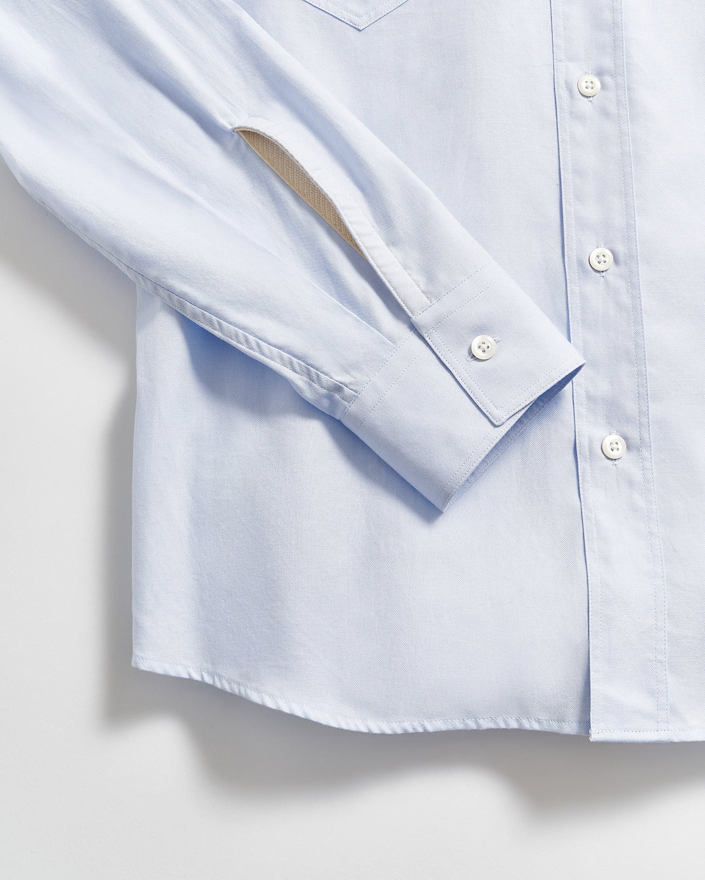 A close-up of a light blue button-up shirt, showing the sleeve, cuff with a button, and part of the front with white buttons against a plain background.