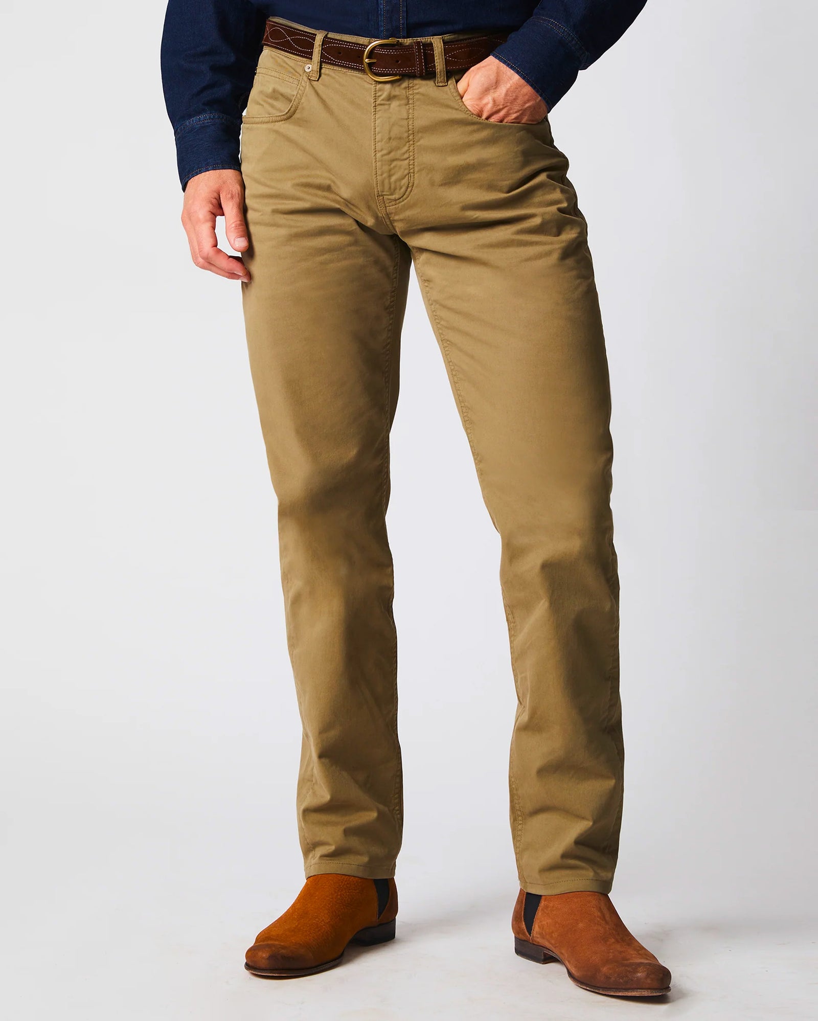 A person wearing tan khaki pants, a dark belt, a dark blue shirt, and brown suede boots, standing against a plain light background. The upper body and head are not visible.