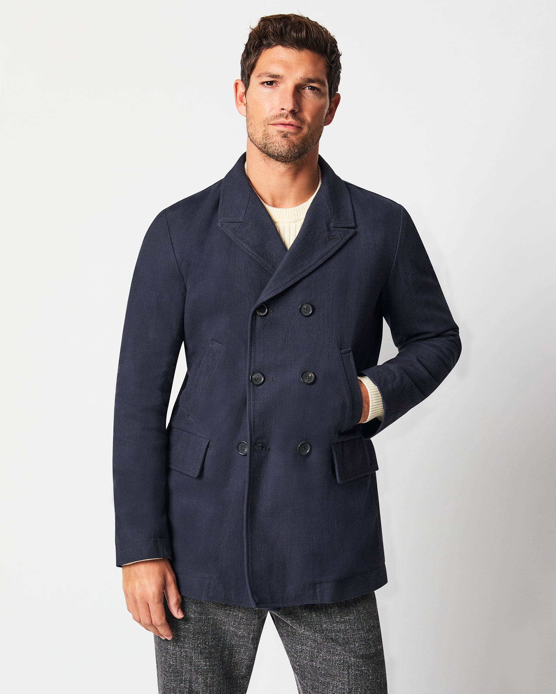 Man wearing a dark blue peacoat and gray trousers standing against a plain background.