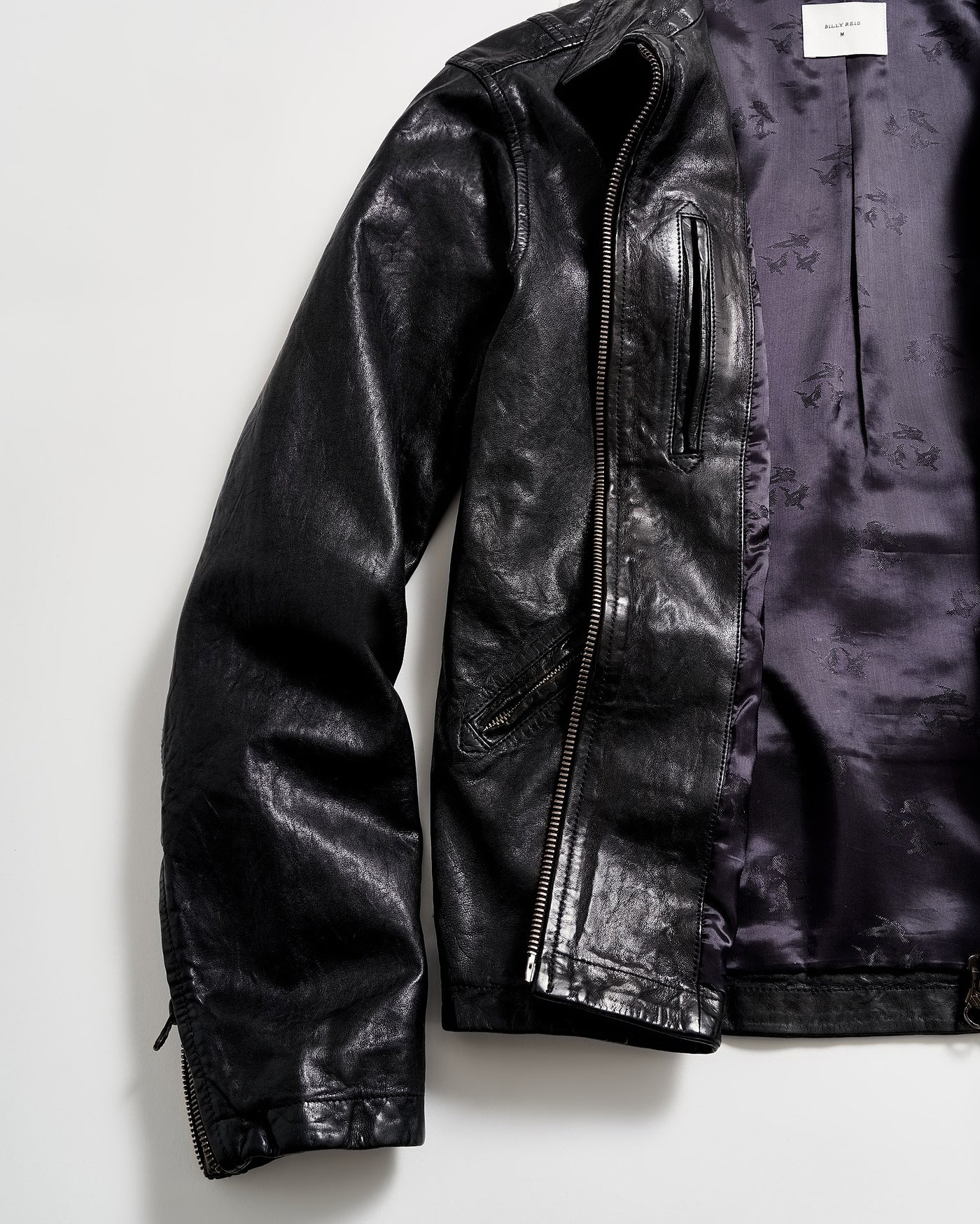 A black leather jacket with a shiny finish is laid flat on a white surface. The jacket is partially unzipped, revealing a purple satin-like inner lining.