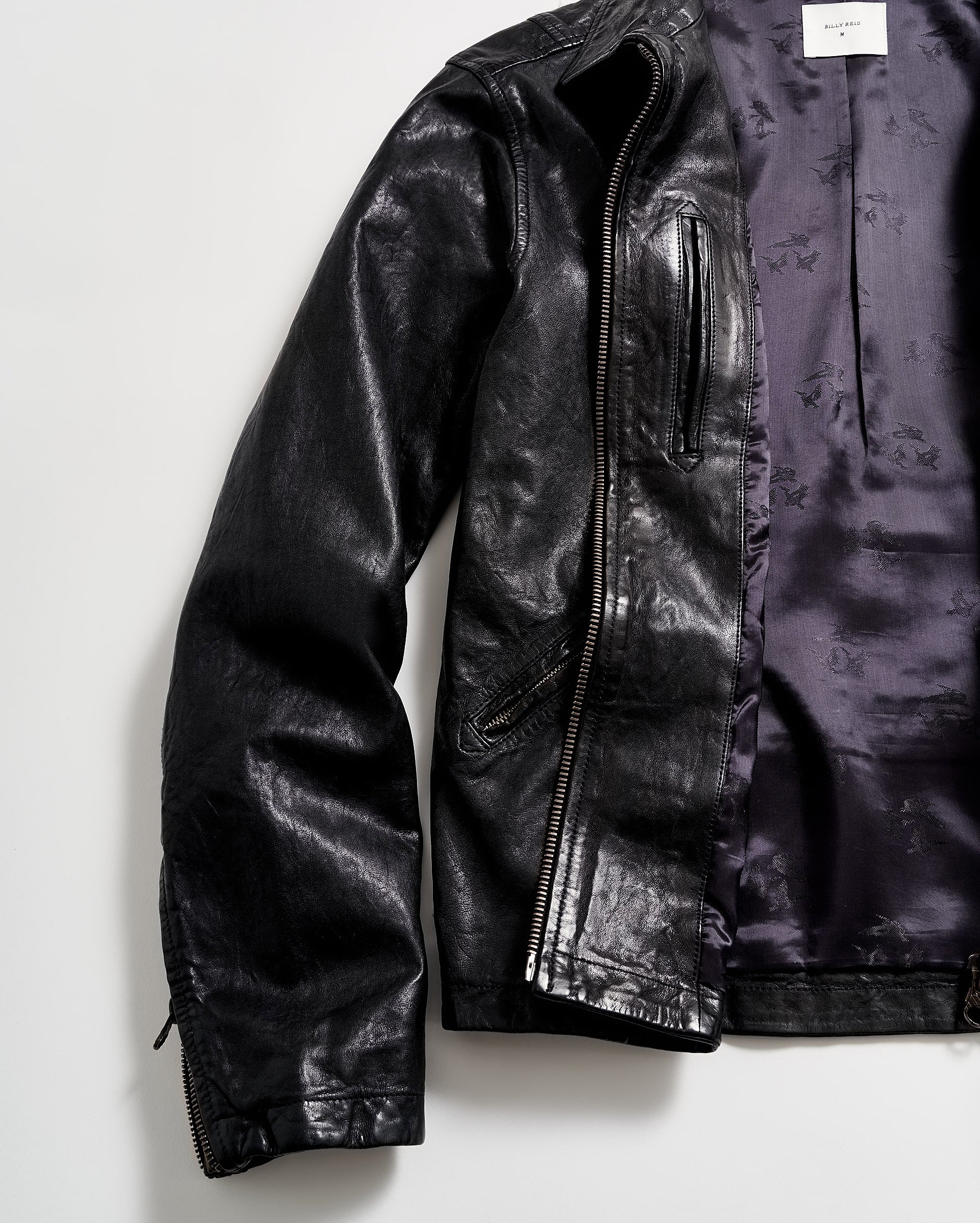 A black leather jacket with a shiny finish is laid flat on a white surface. The jacket is partially unzipped, revealing a purple satin-like inner lining.