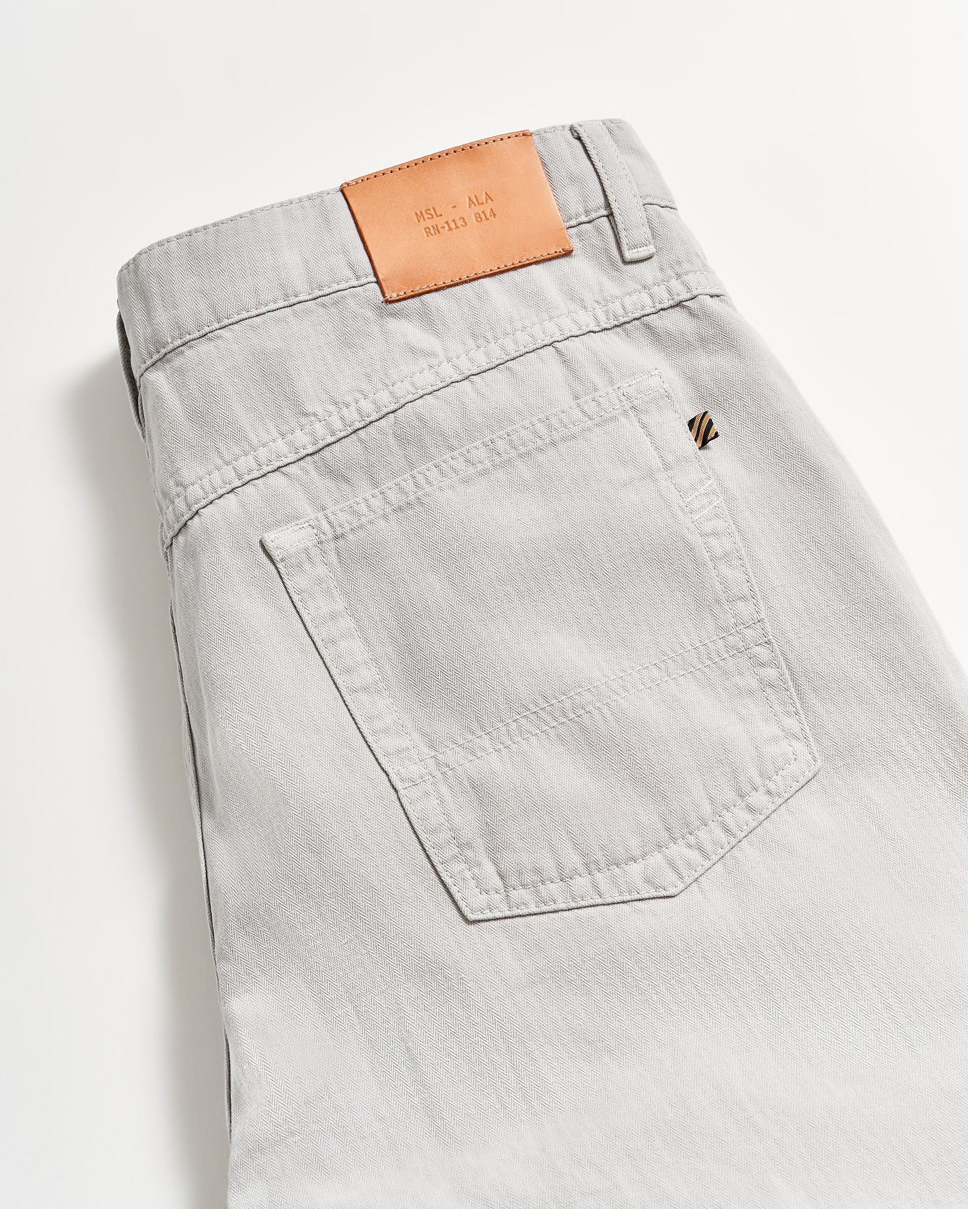A close-up of the back pocket and waistband of light gray jeans with a tan leather patch featuring embossed lettering, laid flat on a white surface.